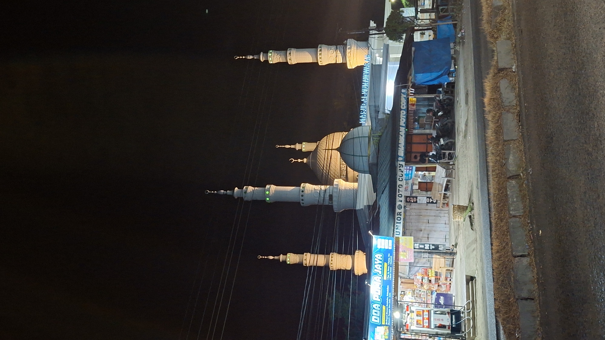 A nighttime, eye-level, full shot captures the Al-Munawwarah mosque in Lhokseumawe, Indonesia, illuminated against a dark sky. The mosque features a large central dome and four tall minarets with pointed tops and decorative banding, some of which are lit with green lights. A sign above the mosque entrance reads "MASJID AL-MUNAWWARAH". In front of the mosque, a row of small businesses lines the street. To the left, a store named "DOA PUMA JAYA" with shelves stocked with goods is visible. Adjacent to it are several other shops, including one with "UNIROR FOTO COPY" and "SHARAKAN FOTO COPY" signage. Several motorcycles are parked in front of these shops. People are seated in front of some of the establishments. The street in the foreground is asphalt, with a grass median bordered by concrete blocks. Electrical wires are strung across the sky.