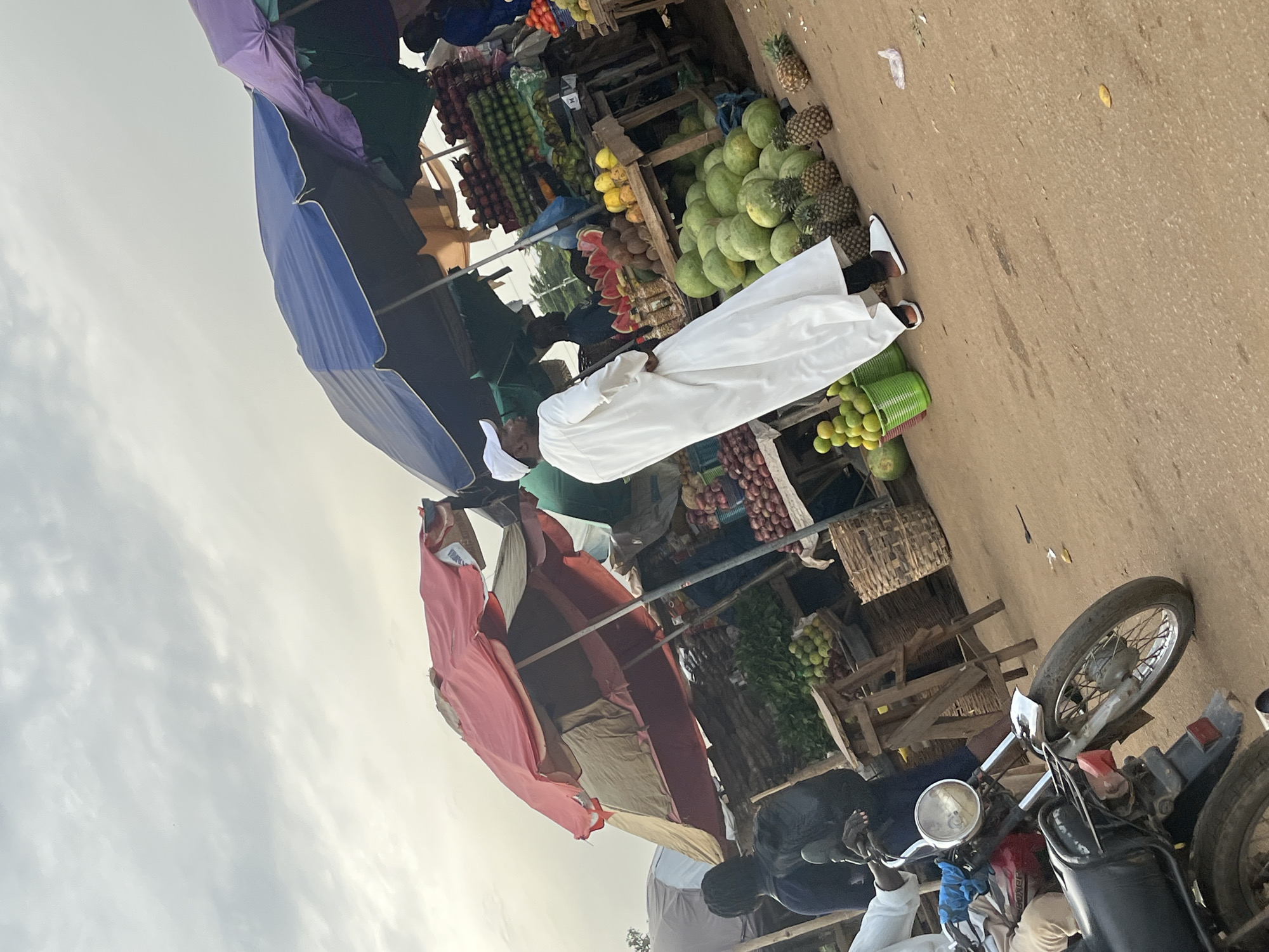 The image depicts an outdoor market scene in Galadimawa Village, Nigeria. A variety of fruits are displayed for sale, including watermelons, pineapples, oranges, and what appear to be onions or similar produce. The market is set up with wooden stalls and various colored umbrellas providing shade.
Several people are present. One individual is dressed in a white thobe and head covering, standing near a display of fruits. Another person is partially visible in a red garment. Further back, more individuals are seen around the stalls, suggesting active commerce. A motorcycle is parked in the foreground, indicating transportation for vendors or customers.
The time of day appears to be late afternoon or early evening, given the quality of light and the warm, earthy tones of the ground. The sky is overcast with some texture, suggesting a partly cloudy day. The setting is a dusty, unpaved market area.
The overall atmosphere seems bustling and typical of a local marketplace. There is no visible text in the image. The focus is on the vibrant display of produce and the daily activities of the people in the village.