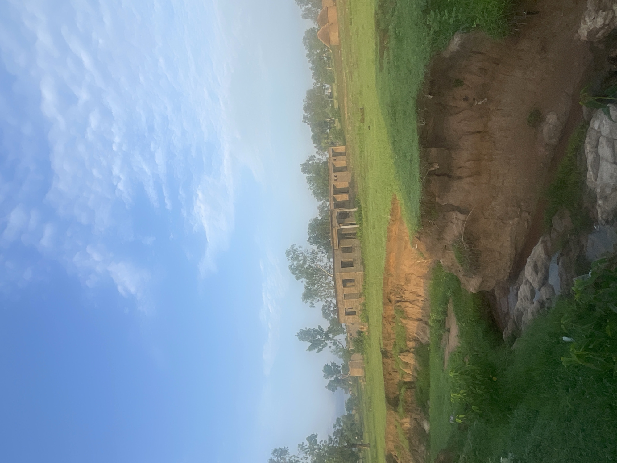 The image depicts a rural landscape with a small stream flowing through eroded earth in the foreground. In the middle ground, there are several unfinished brick buildings and some traditional thatched-roof huts. The background is filled with trees and a hazy sky with some clouds. The scene appears to be located in a remote area, possibly on the outskirts of Jalingo, Nigeria, given the architectural style of the buildings and the vegetation. The lighting suggests it is either early morning or late afternoon. There are no people or animals visible in the image. The overall impression is one of undeveloped or developing rural infrastructure.