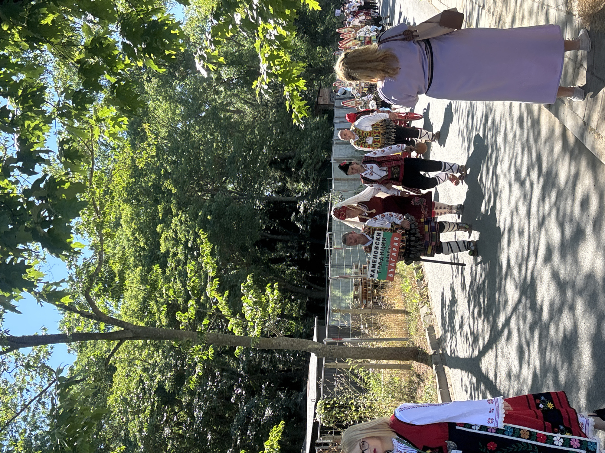 The image depicts a procession of people in traditional Bulgarian costumes walking down a paved street lined with trees. In the foreground, a woman in a red and black embroidered dress with a white blouse stands on the left. To her right, a group of individuals in festive attire, including headwear, vests, and patterned leggings, are walking. One man in the group holds a sign that reads "КАМЕНОВСКИ ТРАДИЦИИ гр. КАМЕНО БЪЛГАРИЯ" which translates to "KAMENOVSKI TRADITIONS town. KAMENO BULGARIA." Further in the background, more participants in similar costumes are visible, as well as a few onlookers. The environment is sunny with dappled shadows from the trees on the pavement. The background shows dense greenery and a fence. The setting appears to be an outdoor event or festival, possibly in the city of Burgas, Bulgaria, given the reference to Bulgaria on the sign.