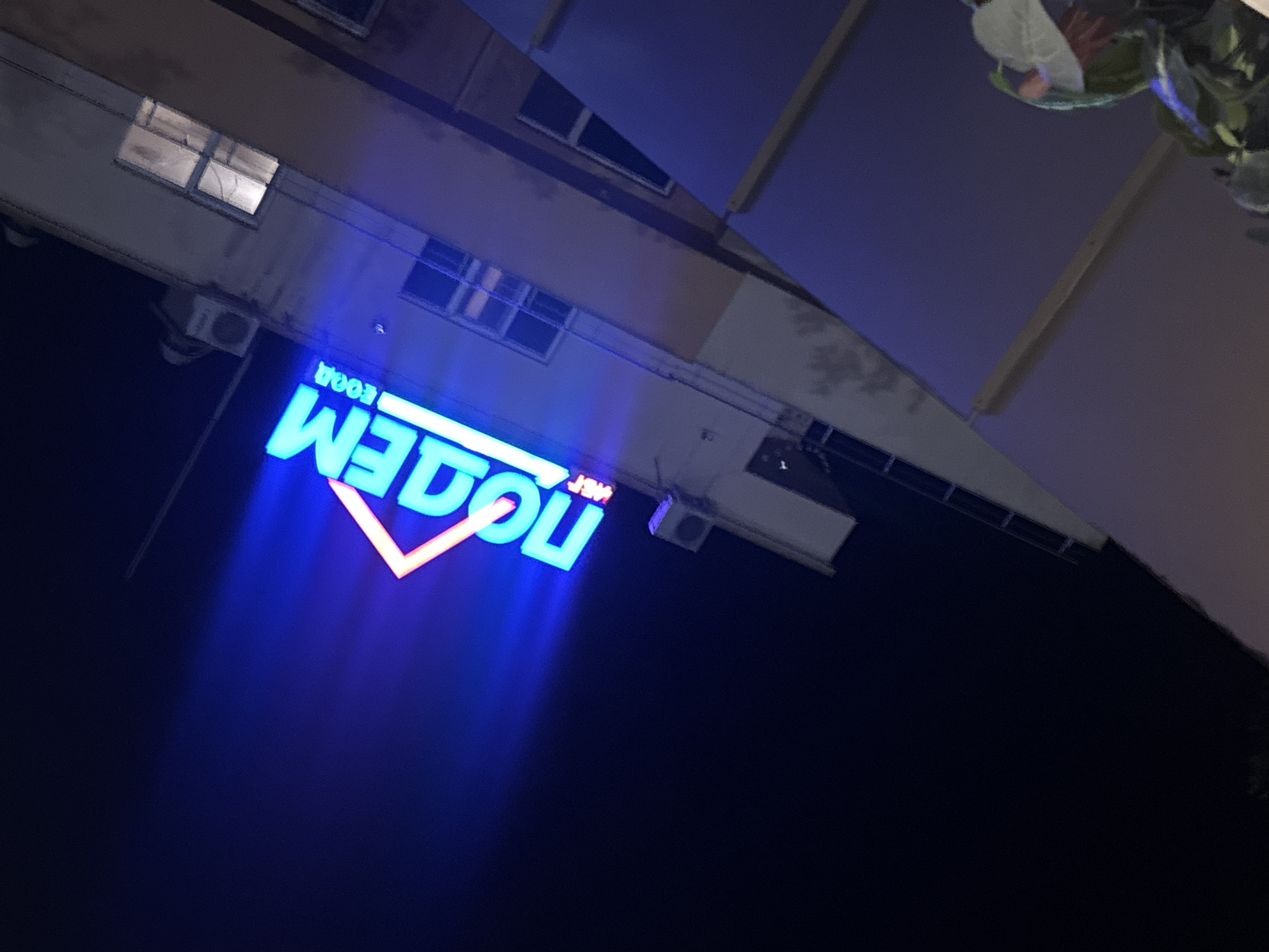 The image shows a neon sign at night, tilted at an angle. The sign, which appears to be in Russian, reads "ПОДЪЕМ FOOD" in bright blue and pink neon letters. The building behind the sign is illuminated and has several windows. The sky is dark and appears to be clear.