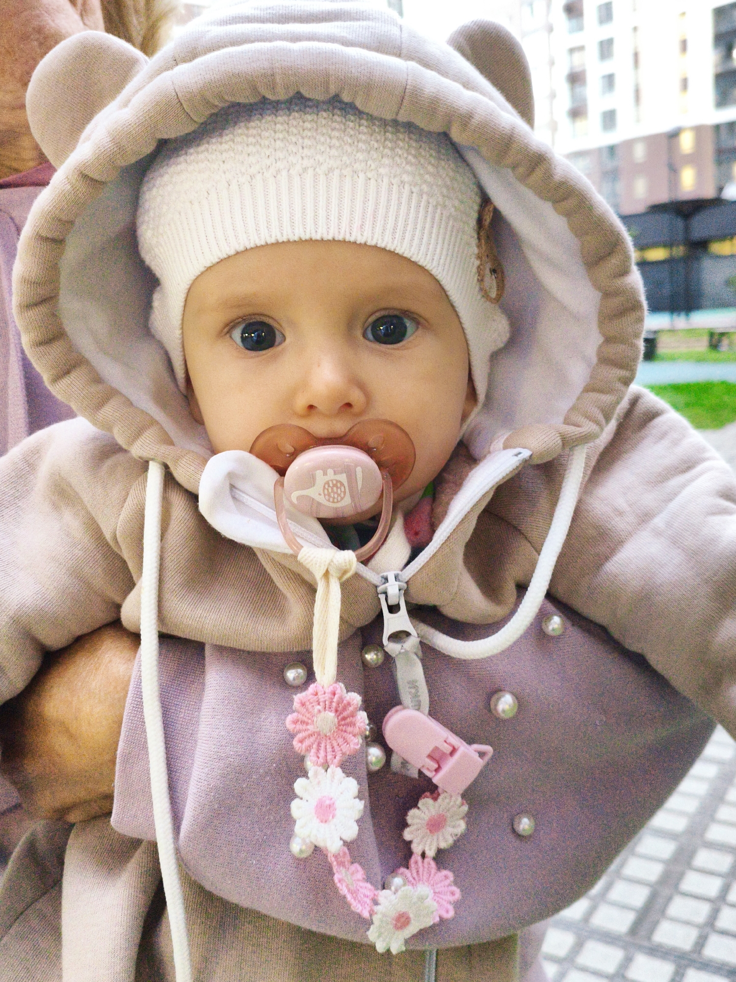 A medium shot captures a baby wearing a hooded jacket with bear ears and a white knitted hat. The baby has big blue eyes and is using a pink pacifier with a white string attached to a pink clip. The jacket is a light brown color with a light purple lining and is adorned with small pearls. A decorative string with pink and white flowers hangs from the zipper. The background shows a blurry urban environment with buildings and a paved walkway.