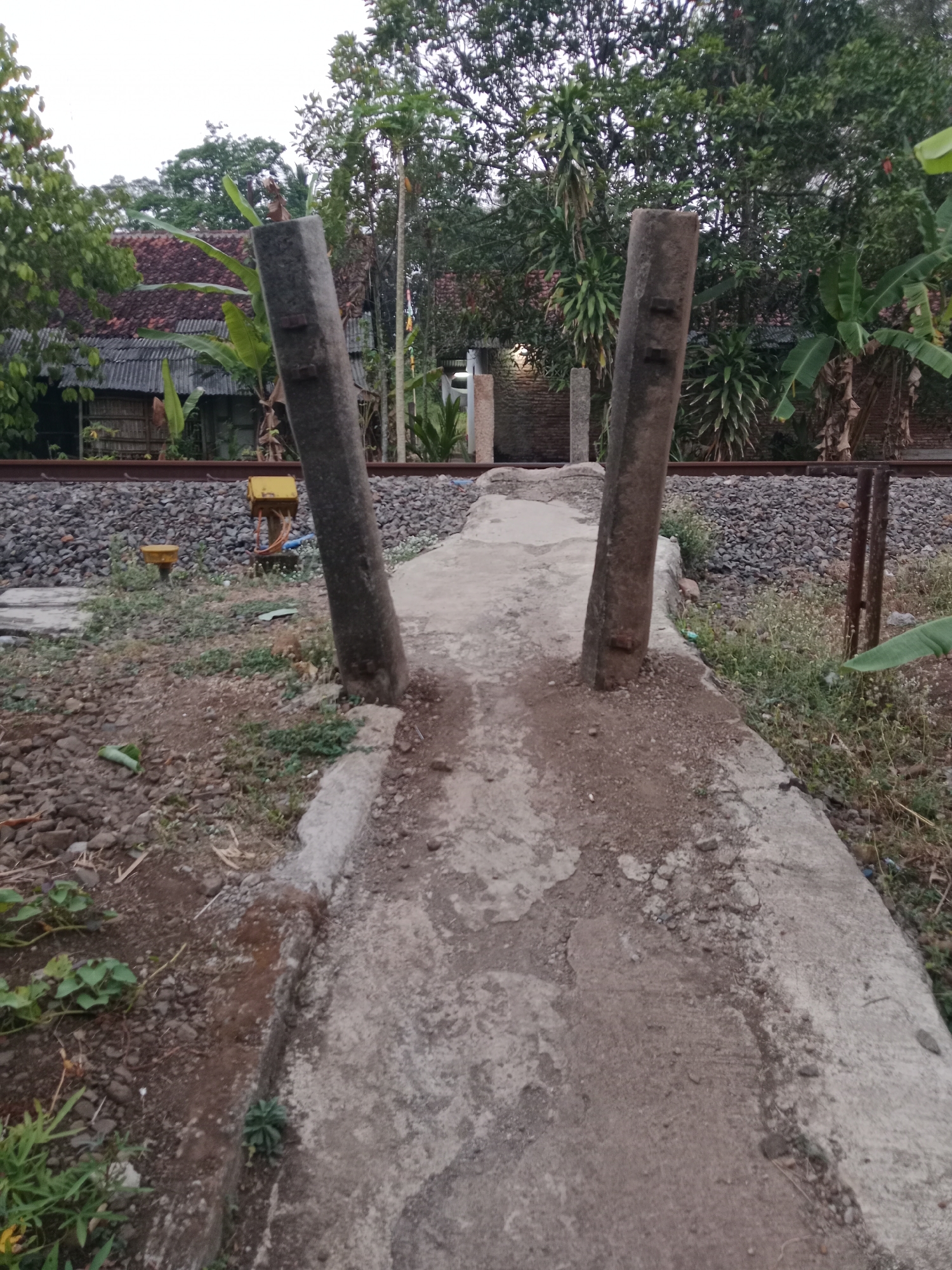 The image shows a concrete path leading towards a railroad track in Bojongmengger, Indonesia. Two weathered concrete pillars, leaning outwards, frame the entrance to the path. Beyond the pillars, the path appears to merge with the ground around the railway. The railroad track, made of gravel and steel, runs horizontally across the middle ground of the image, with a small yellow electrical box visible near the left side. Vegetation, including trees and plants, surrounds the area, with a building featuring a tiled roof visible in the background. The sky is overcast.