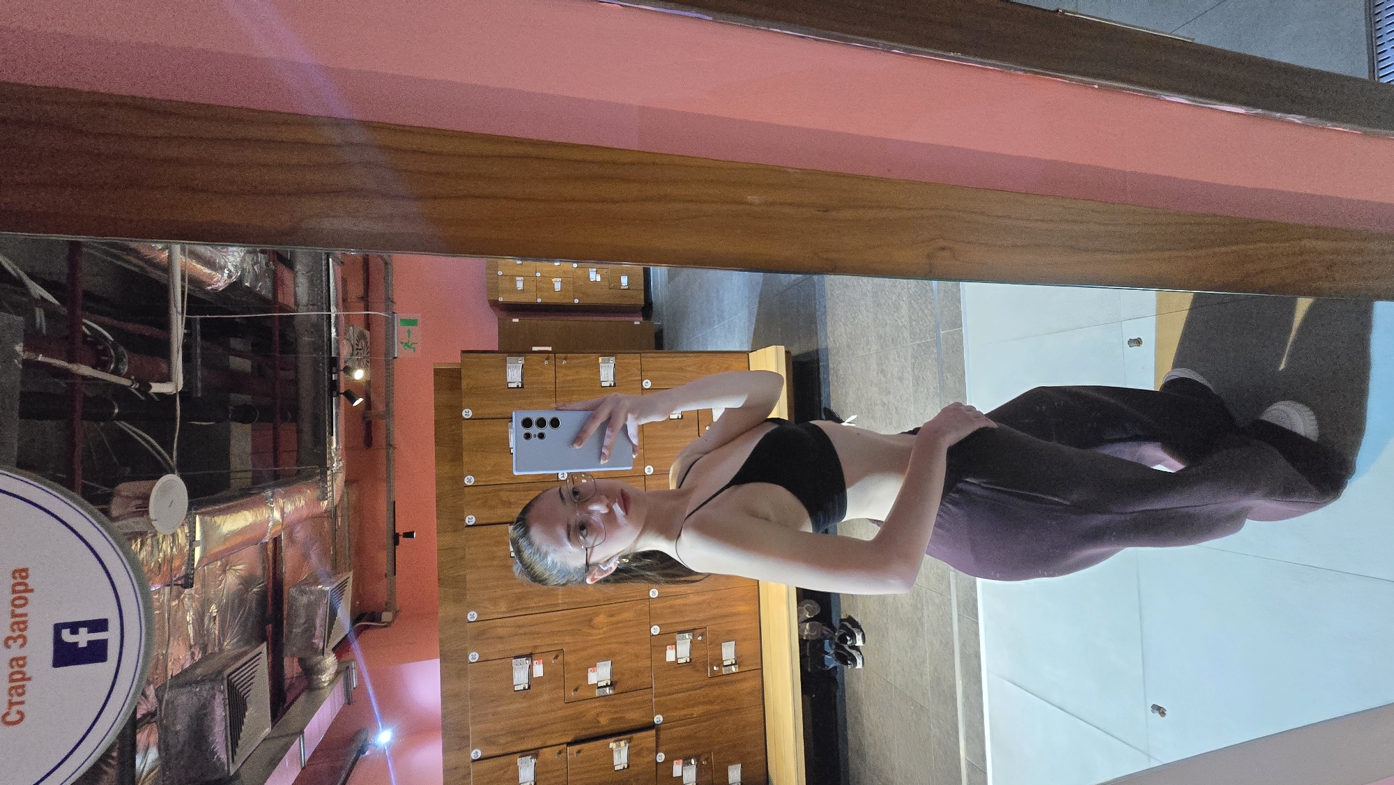 A woman in a black sports bra and dark-colored sweatpants stands in front of a mirrored wall in what appears to be a locker room. She is holding a phone and taking a selfie. Behind her are wooden lockers and some ductwork. A sign with the words "Стара Загора" (Stara Zagora) and a Facebook logo is partially visible. The location is likely a gym or sports facility in the city of Stara Zagora, Bulgaria.