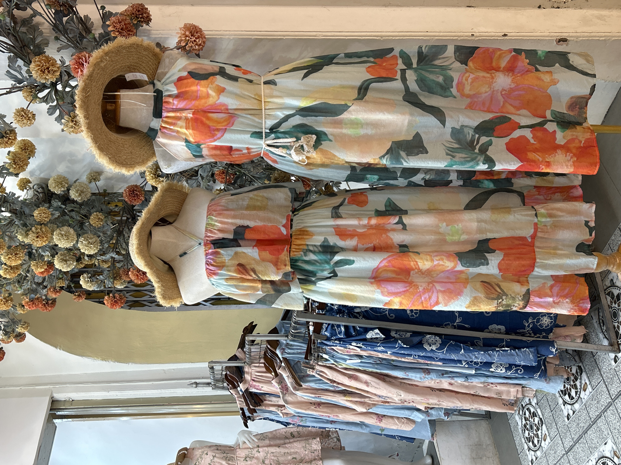 This image depicts the interior of a clothing store, likely located within Phuket City Municipality, Thailand. The scene is dominated by two mannequins adorned with long, flowing floral print dresses. Both dresses feature a halter neckline and a straw hat is placed on each mannequin. The dress on the right has a cream-colored base with large, vibrant orange, pink, and green floral patterns. A thin, braided white belt cinches the waist of this dress, with a small crocheted detail hanging from it. The dress on the left, partially visible, appears to have a similar floral print and is strapless.
In the background, to the left, a rack of clothing is visible, featuring an assortment of pastel-colored dresses and tops with floral or botanical prints in shades of pink and blue. Further left, a portion of another mannequin wearing a light pink dress is visible through a doorway.
The store's decor includes a large, decorative floral arrangement hanging on the wall behind the mannequins. This arrangement consists of numerous textured, globe-shaped flowers in shades of cream, tan, and burnt orange, interspersed with dark green leaves. The floor is tiled with a patterned design featuring circular motifs. The overall lighting suggests an indoor retail environment, possibly illuminated by natural light from the adjacent doorway and ambient store lighting. The aesthetic suggests a boutique specializing in summery or resort wear.