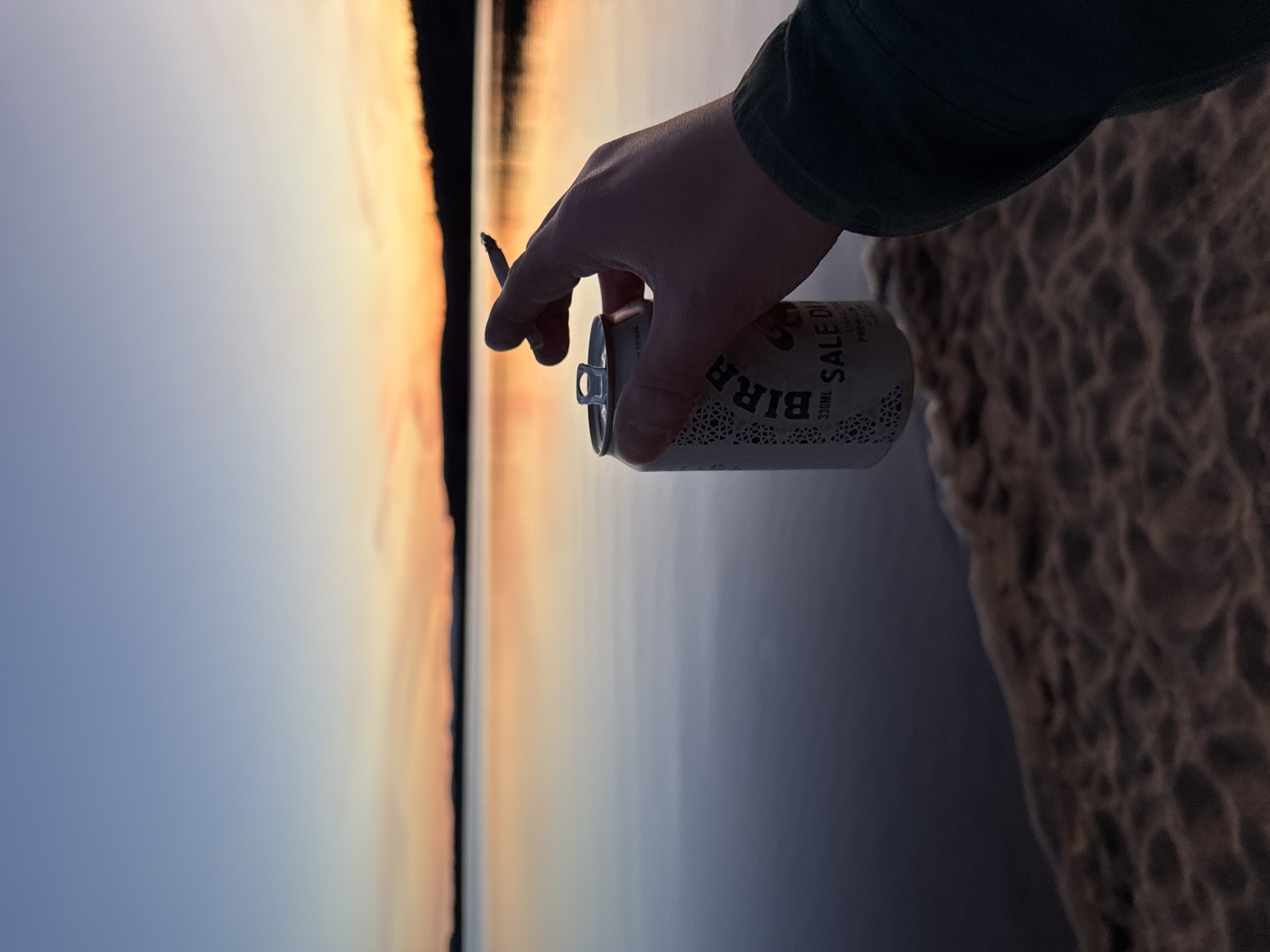 The image, viewed in landscape orientation, captures a person's right hand holding a lit cigarette and a beverage can, silhouetted against a scenic background. The hand and part of a dark green sleeve are in the mid-ground. The background features a serene body of water reflecting a vibrant sunset or sunrise sky, with hues of orange, yellow, and soft blue meeting a distant, dark horizon line. The water appears calm, mirroring the sky's colors. To the right, a textured, uneven surface, likely sand or dry earth, fills the foreground.
The person holds a lit cigarette between their index and middle fingers. The beverage can is light-colored, possibly white or cream, with a subtle pattern and dark text. Visible words on the can include "BIRRA" prominently, and "SALE DI" and "330ML" beneath it, suggesting it's a can of beer. The pull tab of the can is open. The scene evokes a moment of relaxation by a lake or sea during the golden hour.