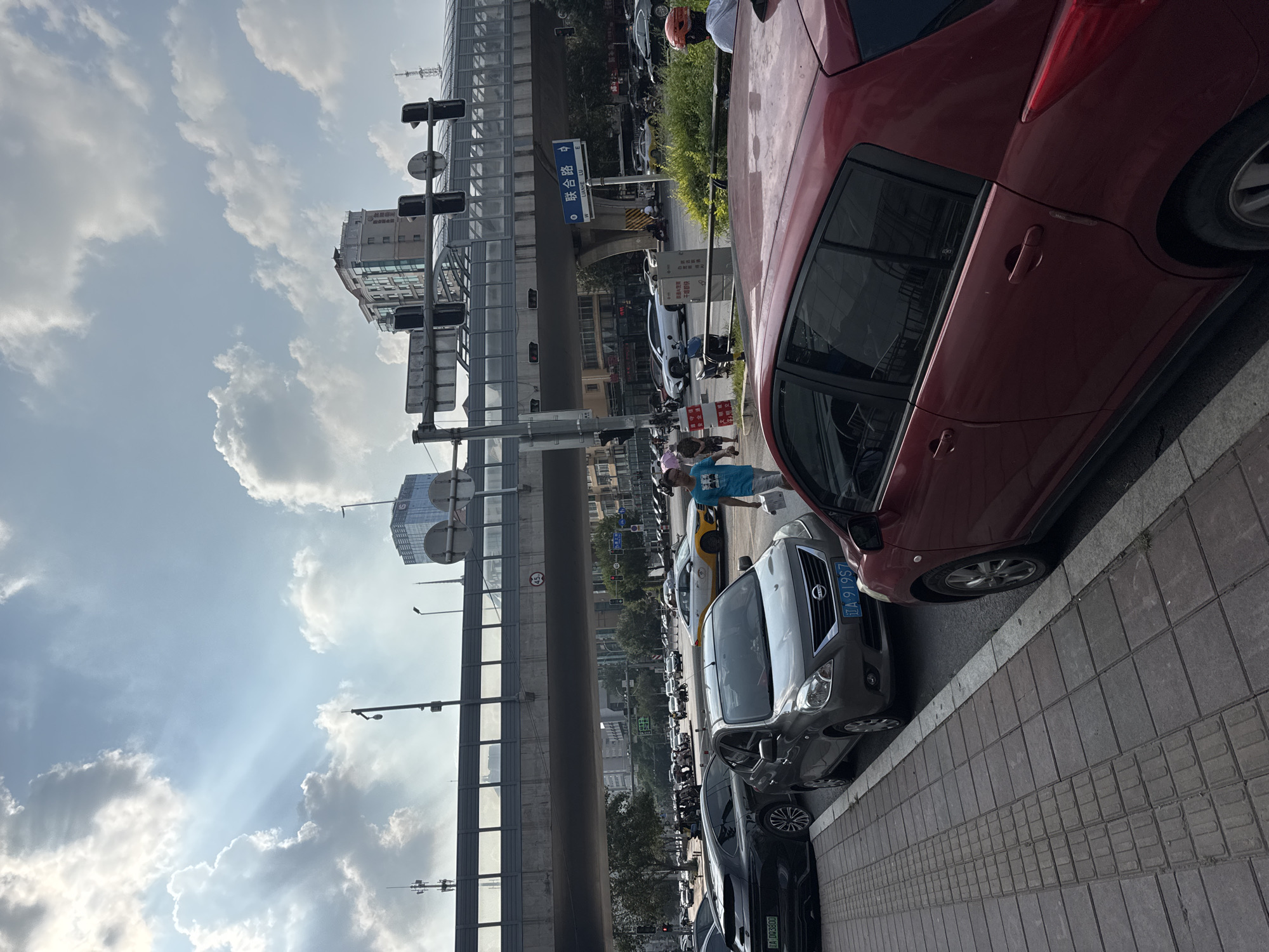 This image captures a street scene in Dadong District, China, on a sunny day with scattered clouds. A busy road is visible, with several cars parked on the side and others in motion. Traffic lights and street signs indicate an urban environment. An elevated walkway or overpass spans across the road, with buildings visible in the background. Several pedestrians are present, including a person walking on the sidewalk and others in the street. The overall impression is one of an active city intersection.