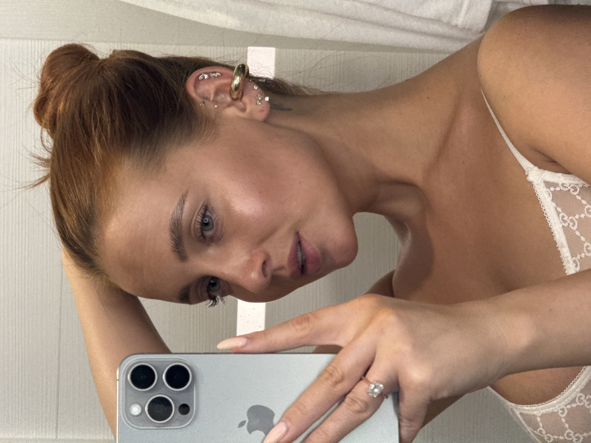 In a mirror selfie, a young woman with auburn hair pulled into a high bun is shown from the chest up. She is holding a silver smartphone in her left hand, using it to take the picture. Her left arm is bent, with her hand resting on the back of her head. She is wearing a white lace bra. She has multiple earrings in her left ear, including a large gold cuff and several smaller stud earrings. A diamond ring is visible on her left ring finger. The background appears to be a bathroom, with a tiled wall and a white towel hanging on the right. The lighting is bright and even. There is no indication that this image was taken in Chaikovsky, Russia.