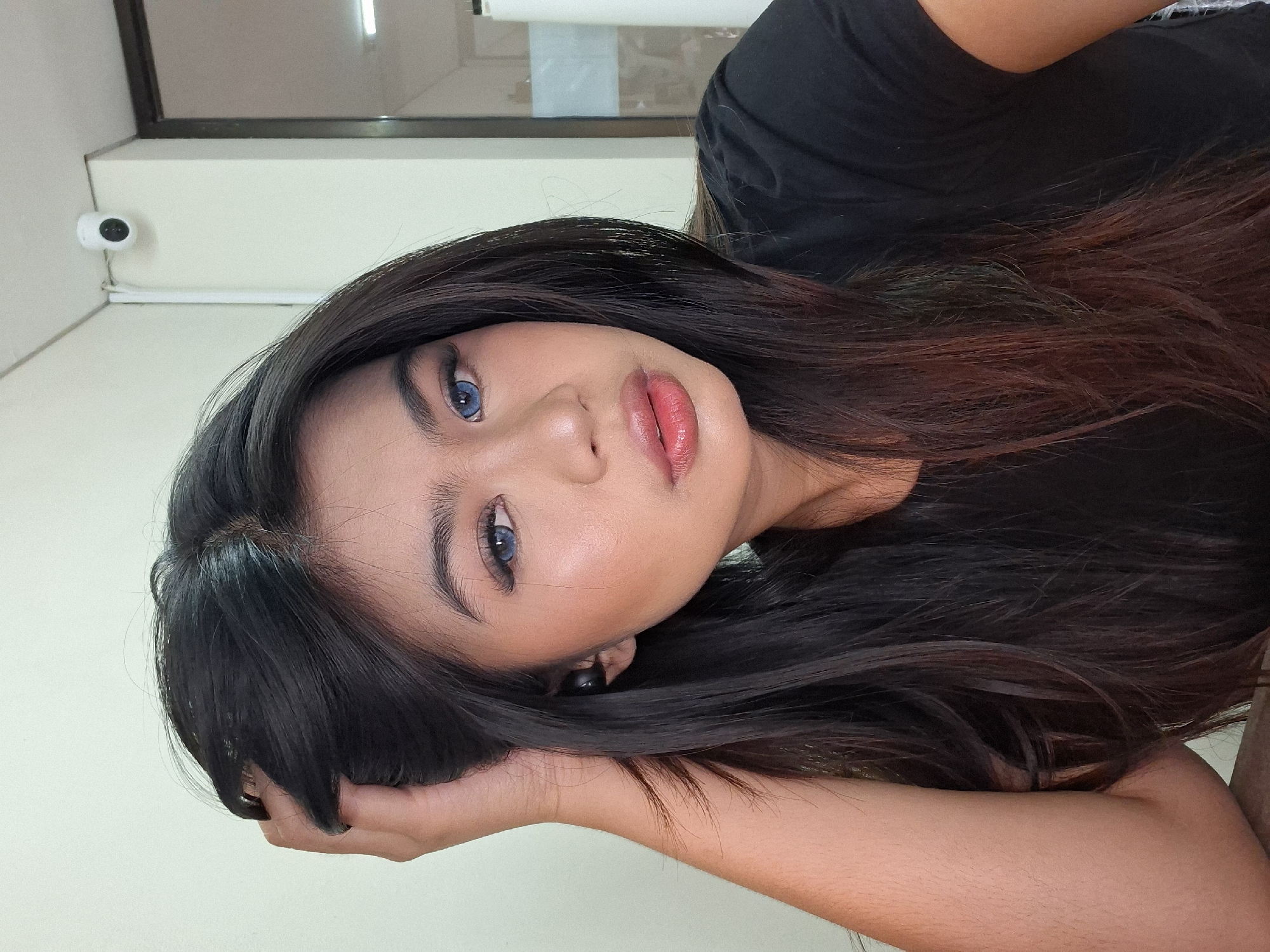 A young woman with dark hair and blue contact lenses is captured in a close-up, waist-up photograph. She is indoors, likely in an office setting, given the visible security camera mounted on a white wall in the background. The woman is wearing a black t-shirt and has her left hand raised, touching her hair. She is also wearing black earbuds in her left ear. The lighting appears to be artificial and the background is a plain, light-colored wall. The image is a selfie, with the camera focused on her face and upper body. The overall impression is one of casual pose and direct engagement with the camera.