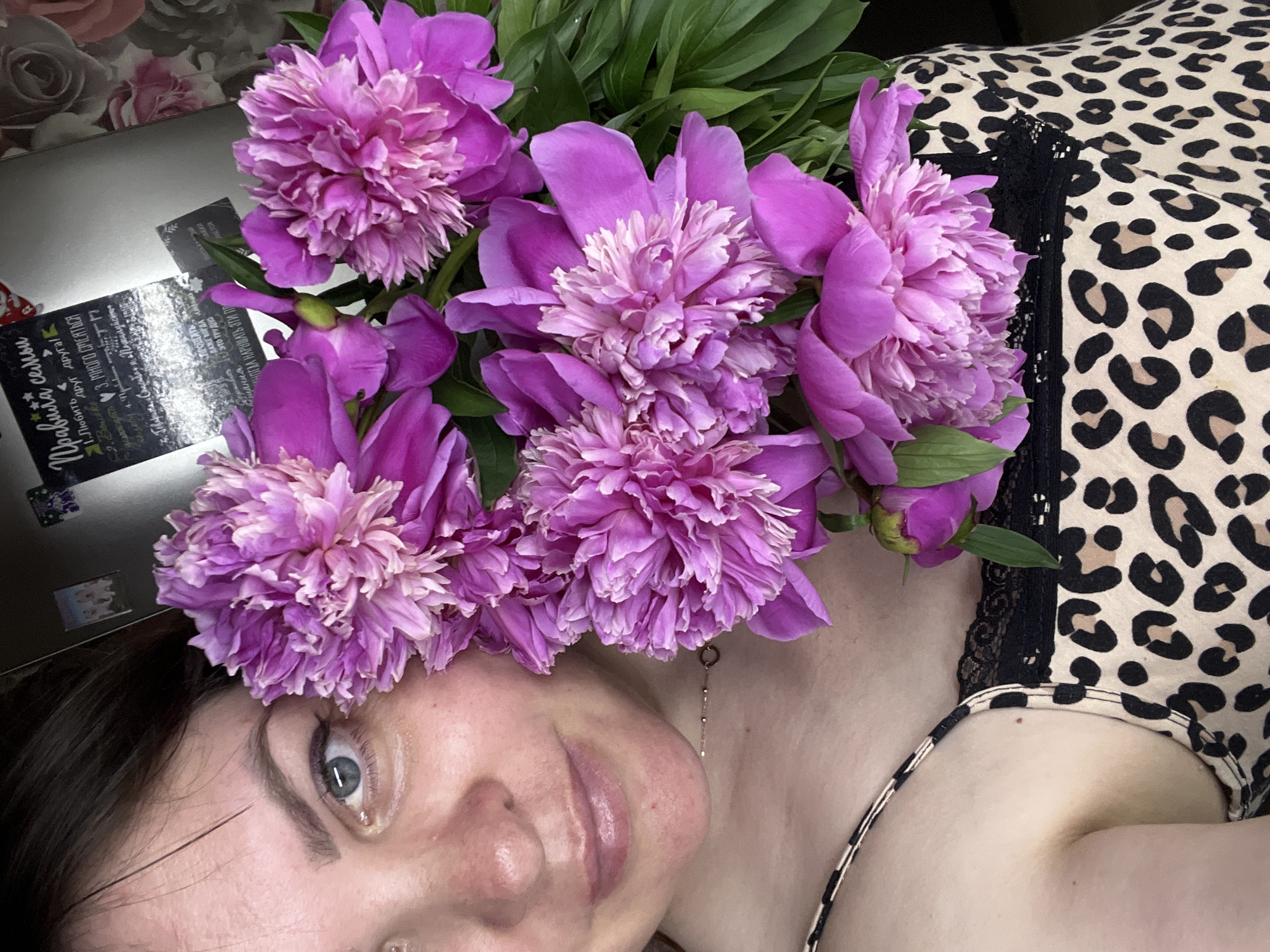 The image is a close-up, selfie-style photograph featuring a woman holding a bouquet of pink peonies. She has dark hair, blue eyes, and fair skin with some freckles. She is wearing a leopard-print camisole with black lace trim. The background appears to be a kitchen, with a refrigerator visible on the right side of the image, adorned with various magnets and stickers. One sticker contains text that appears to be in Russian. There are no discernible elements that specifically place the image in Noviy, Russia. The lighting suggests an indoor setting, likely during the daytime.