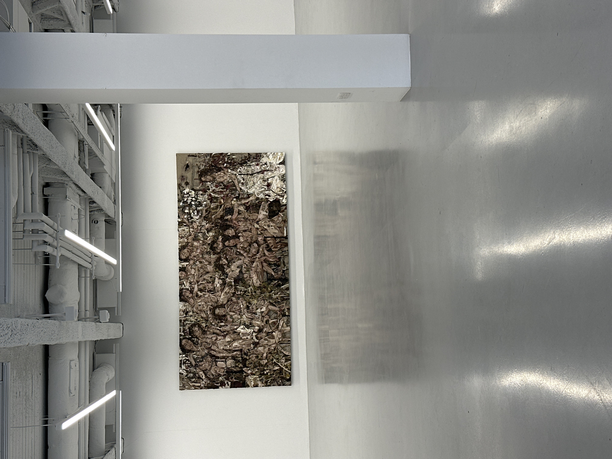 The photograph depicts the interior of a modern art gallery, possibly in London, United Kingdom, suggested by the architectural style and the minimalist aesthetic commonly found in contemporary art spaces in the city.
The space is dominated by white surfaces, including the walls, ceiling, support beams, and the floor, which reflects the overhead lighting, creating bright highlights. A network of painted white pipes and conduits runs along the upper left side of the image, adding an industrial feel to the otherwise clean and sterile environment.
On the wall, an abstract artwork is displayed. It is characterized by a dense and intricate pattern of earth tones, possibly depicting a complex landscape or organic forms. Its composition is non-representational, encouraging subjective interpretation.
The scene is devoid of people. The photograph emphasizes the interplay of light and shadow on the pristine surfaces and the contrast between the functional elements of the architecture and the artistic piece on display.