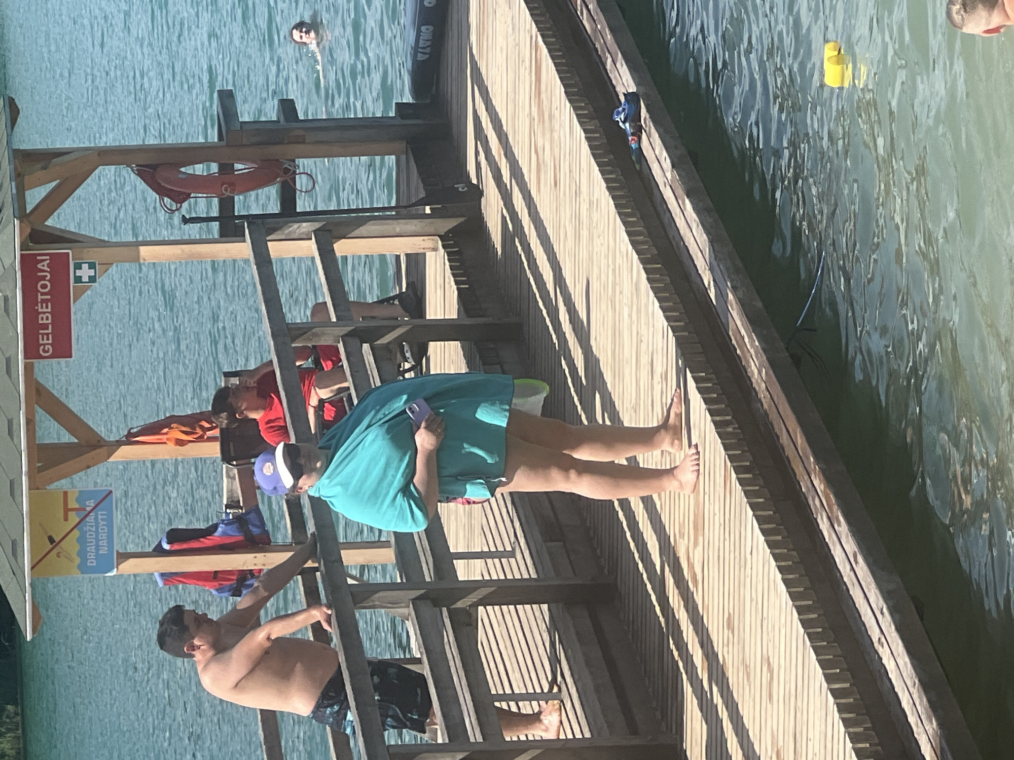 This image depicts a sunny day at a lake or waterfront in Vilnius, Lithuania. The scene is centered around a wooden pier extending into clear blue water. Several people are present on and around the pier. In the background, a person is swimming in the water. On the pier, a person wearing a blue hat and a teal cover-up is holding a phone. Another person is climbing a ladder on the pier, and a third person is shirtless, wearing patterned shorts, and appears to be resting or preparing to enter the water.
Objects visible include a life preserver hanging on the pier structure, a paddleboard near the edge of the pier, and two yellow buoys floating in the water. The pier itself is constructed of light-colored wooden planks, casting long shadows that indicate it is likely midday or early afternoon. The water is calm with gentle ripples.
There is visible text on a red sign affixed to the pier structure that reads "GELBĖTOJAI", which translates to "Lifeguards" in Lithuanian. Below this, another sign with yellow and blue elements displays a pictogram of a person diving and the text "DRAUDŽIAMA NARDYTI", meaning "Diving prohibited". The overall atmosphere seems relaxed and recreational, with people enjoying a day by the water.