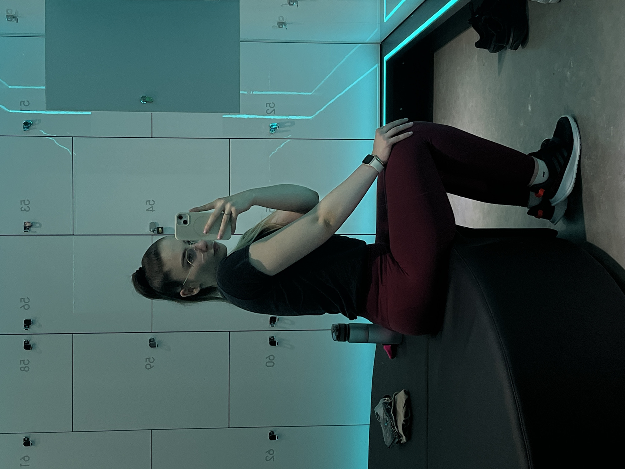 A woman is sitting on a black, curved bench in a room with gray lockers, taking a selfie with her smartphone. She has fair skin, dark hair in a ponytail, and wears glasses. She is dressed in a black t-shirt and maroon leggings. A black athletic shoe is on her right foot. A water bottle and other items are also on the bench next to her. The locker room is lit with a bluish light. The location is Unknown, Unknown.