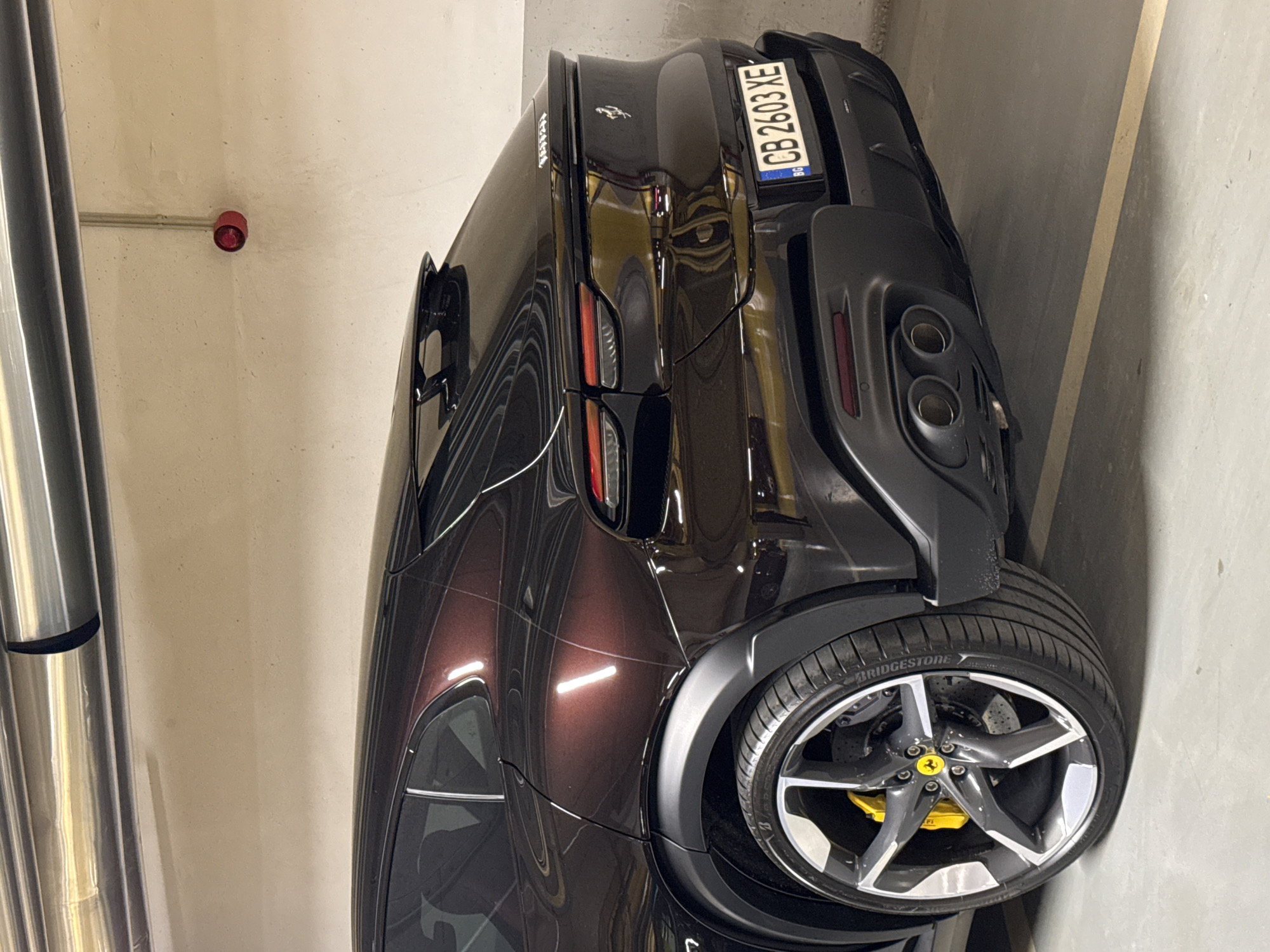 The image depicts the rear portion of a dark brown or black Ferrari parked in what appears to be an underground parking garage. The car is positioned close to a concrete wall. The vehicle's license plate reads "CB 2603 XE" with the Bulgarian flag indicated on the left side. The tire has "BRIDGESTONE" printed on the side. The wheel rim is silver with a yellow Ferrari logo in the center. A red fire alarm or sprinkler system component is visible on the ceiling. Metal pipes are also visible on the left. Based on the license plate, the location is in Bulgaria, specifically Sofia.