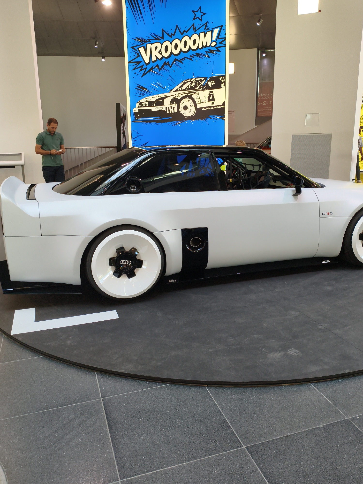 A white Audi rally car is on display indoors, likely at the Audi museum mobile in Ingolstadt, Germany. The car is positioned on a dark circular platform with a white directional arrow painted on it. The car features a large, white, futuristic wheel with a black Audi emblem in the center. A man in a green polo shirt and dark pants is standing to the left of the car, looking down at his phone. Behind the car, a large blue graphic with the word "VROOOOM!" in white lettering and a cartoon depiction of a rally car is displayed on a wall. The environment appears to be a museum or exhibition space, with concrete floors and modern architectural elements.
