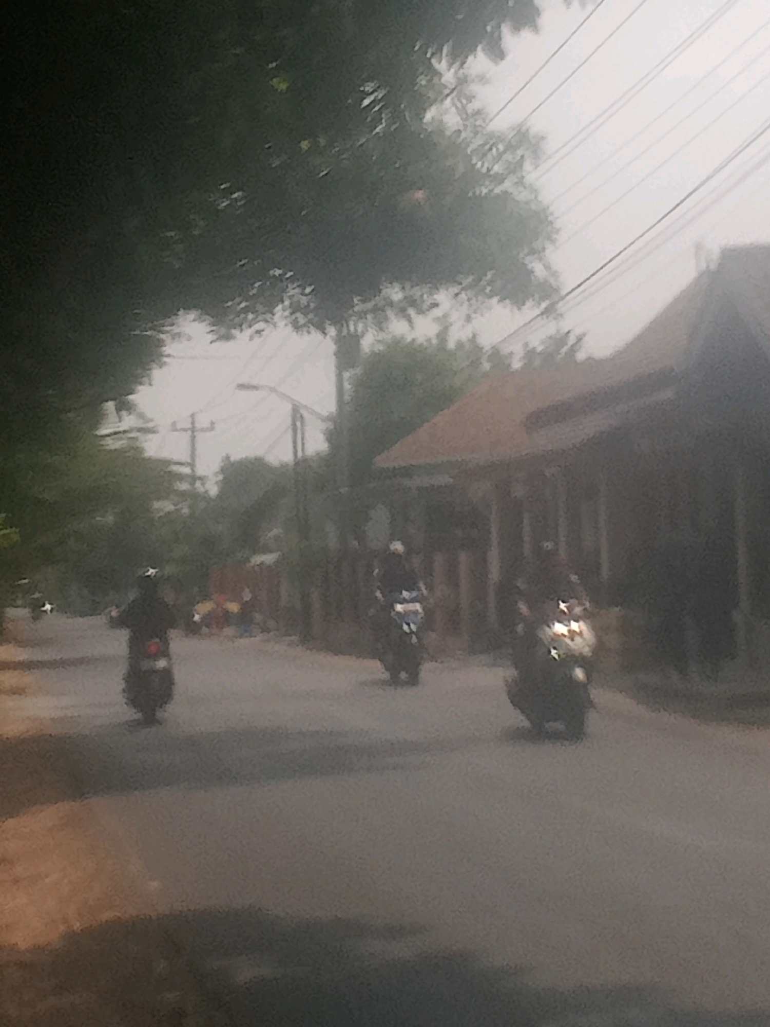 The image depicts a street scene in Lebakgowah, Indonesia. In the foreground, the asphalt road is visible, with a tree casting a shadow on the left side. Three motorcycles are present on the road. The first motorcycle, on the left, has a rider wearing a dark jacket and a white helmet, moving away from the viewer. The second motorcycle is in the middle of the road, with a rider wearing a dark helmet and jacket, also moving away. The third motorcycle is on the right, with its headlight illuminated, and the rider is wearing a helmet and dark clothing, facing the viewer. In the background, there are buildings with brown roofs and trees lining the street. Power lines are visible in the sky. The overall atmosphere is hazy and overcast.