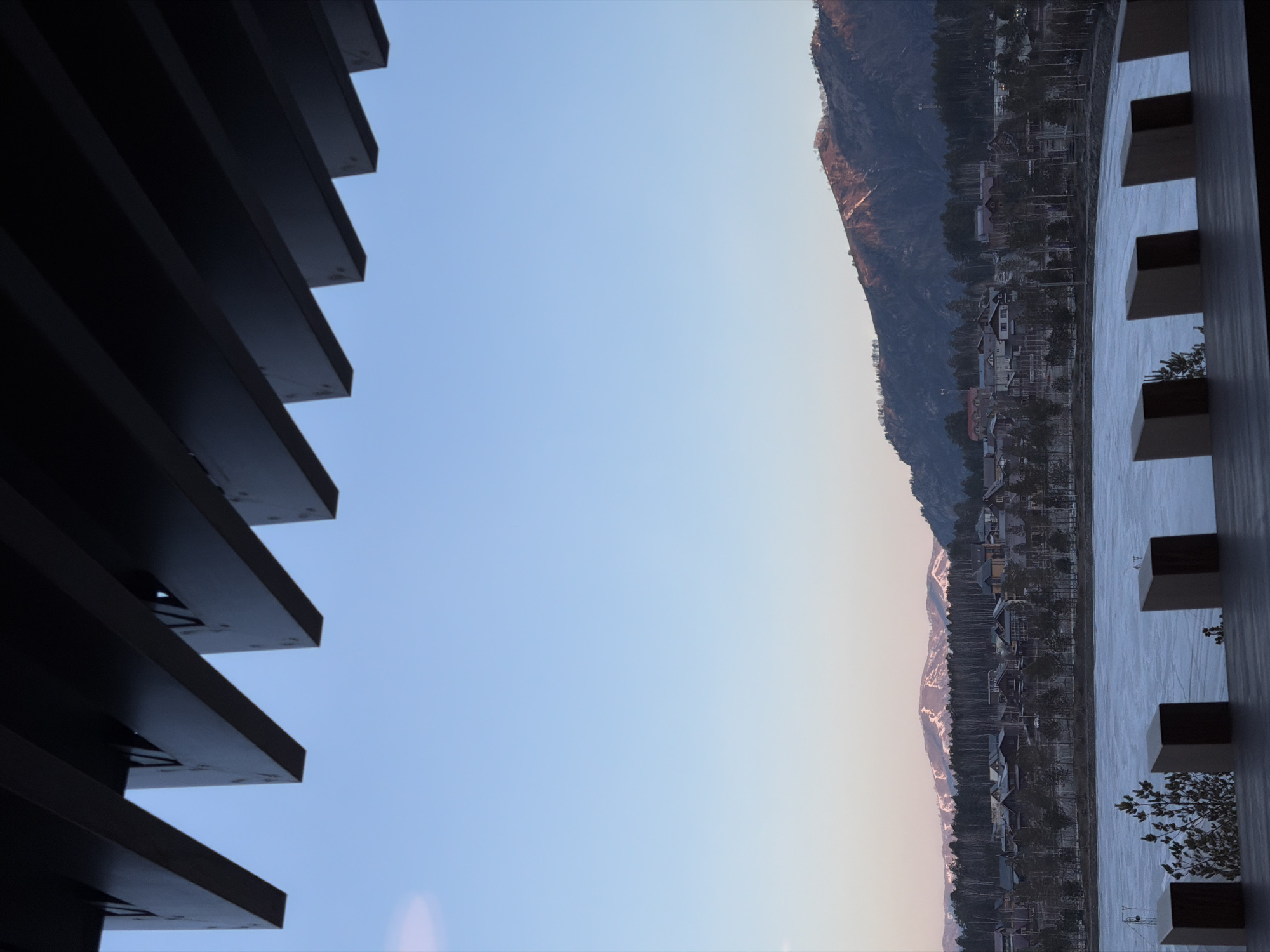 The image, oriented vertically, shows a view from an elevated position in Озёрное, Russia. A linear mountain range with a mix of vegetation and sparse snow cover dominates the right side of the frame. A residential area with clustered buildings is nestled at the base of the mountain. On the far left is the dark structure of a multi-layered building, with horizontal ledges running at an angle toward the top-right. Along the right edge of the image, a building exterior with alternating square projections is visible. The sky occupies the central portion of the frame, transitioning from a pale blue to a lighter, almost pastel hue closer to the horizon.