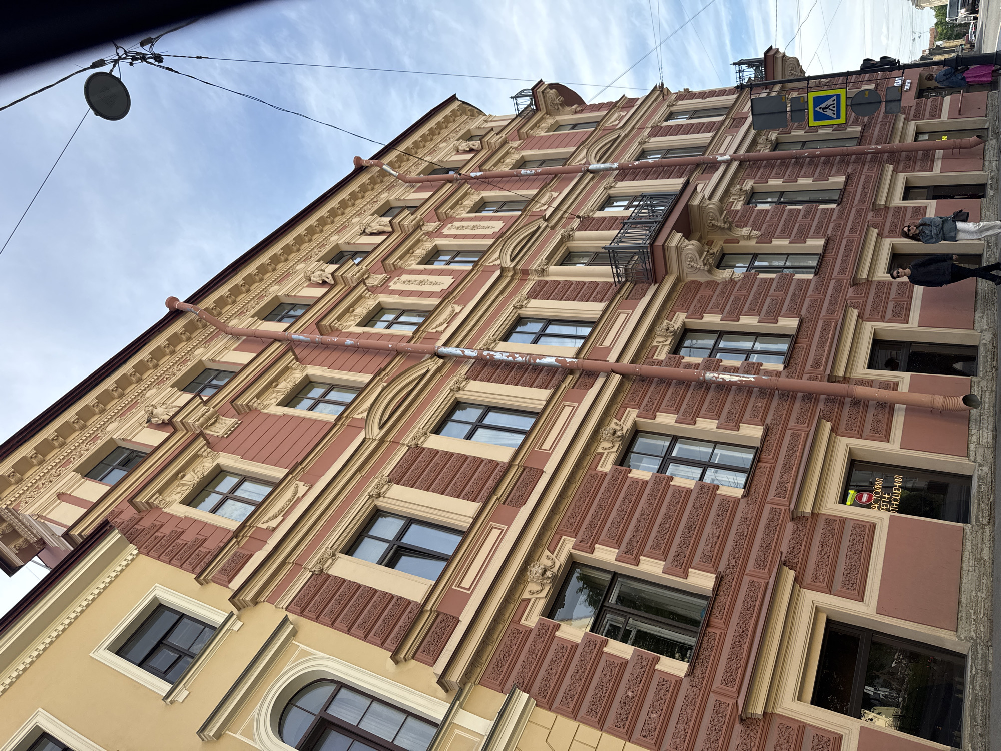 This media shows a street scene in Saint Petersburg, Russia, captured from a low angle looking upwards at the facade of a multi-story building. The building features an ornate architectural style, characterized by a primary color scheme of terracotta red/pink and light yellow/beige.
The facade is rich in decorative elements, including elaborate relief carvings that adorn the wall panels and frame the numerous windows. The windows, mostly rectangular with dark frames, are set within contrasting light yellow/beige surrounds. Several weathered, reddish pipes run vertically and horizontally across the building's exterior, displaying some signs of peeling paint. A small, dark-colored metal balcony or decorative structure is visible on one of the upper floors.
At the street level, several individuals are seen walking on the sidewalk. A yellow and blue pedestrian crossing sign is prominently displayed on a pole. On a ground-floor window, Cyrillic text reads "НАСТОЯЩИЙ ВЕТЕРАН ОТНОШЕНИЙ" (Real Veteran of Relationships), and a partial sign for "ТАКСИ" (Taxi) is also visible.
The sky above is bright and partly cloudy, indicating daytime with fair weather. Overhead, power lines and a circular street lamp are noticeable. The unique architectural style, the Cyrillic text, and the urban setting strongly suggest the location is Saint Petersburg, Russia.
