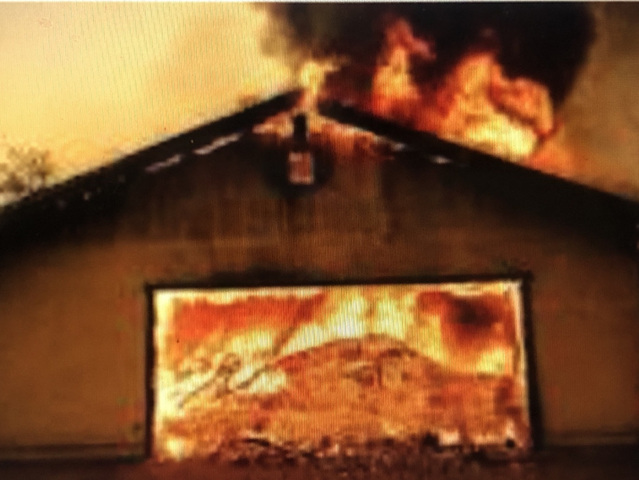 This image depicts a structure in Kafr Sousa Municipality, Syria, actively engulfed in fire. On the left side, a rectangular opening, consistent with a window or framed aperture, is filled entirely with intense orange and yellow flames, indicating an internal blaze. To the right, a darker structural element, possibly a wall or beam, is visible, with more flames and dark smoke billowing from beneath and around it. A diagonal dark line, likely representing a roofline or architectural feature, separates areas of fire and smoke. The scene is dominated by the bright, luminous glow of the fire, casting warm tones across the visible surfaces, with no discernible individuals or explicit actions beyond the ongoing combustion.