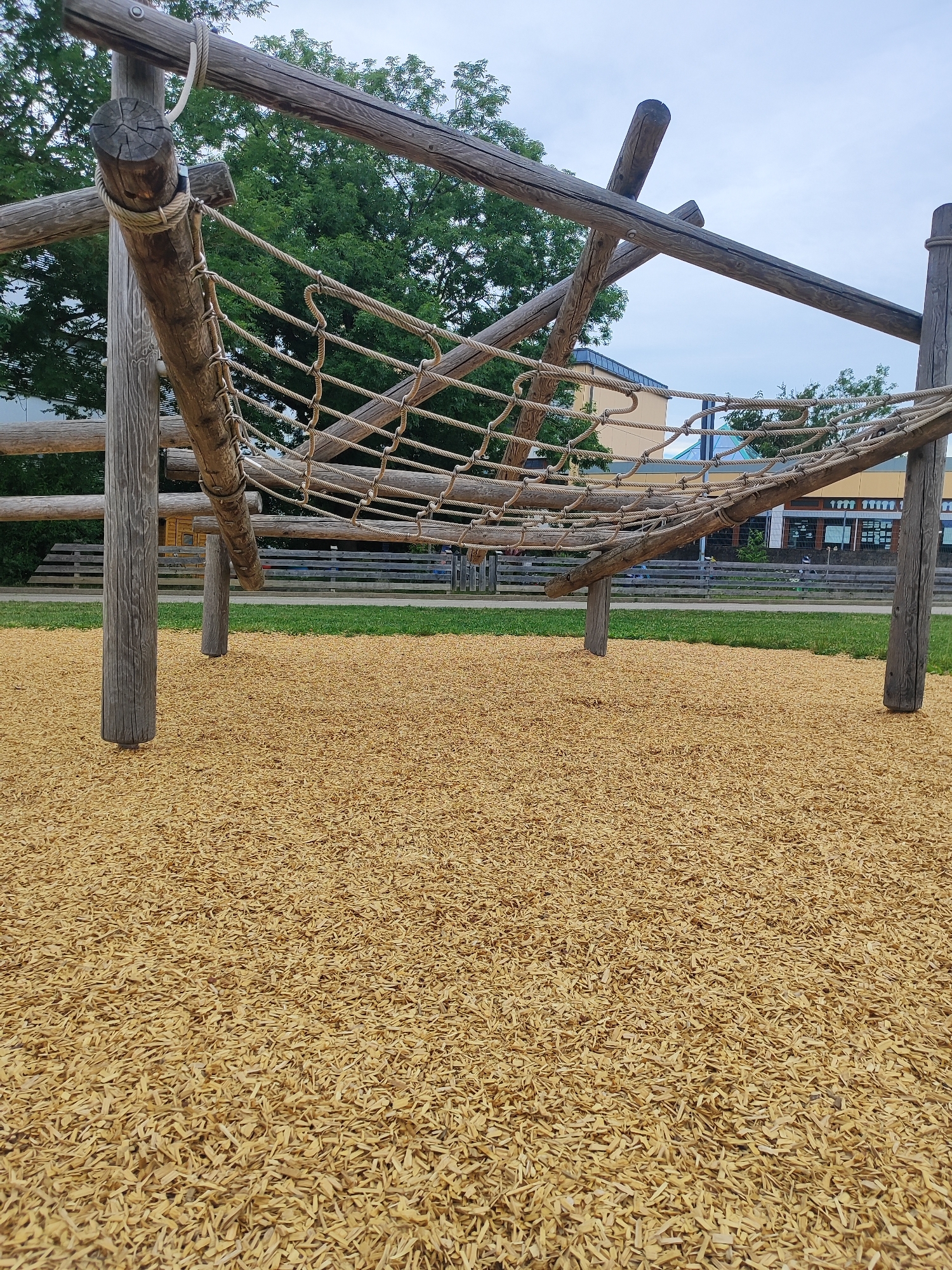 The image depicts an outdoor playground structure in Diedorf, Germany. The structure is made of large, rough-hewn wooden logs forming an angled frame from which a thick rope net is suspended. The net appears to be a climbing or balancing element. The ground beneath and around the structure is covered with a thick layer of light-brown wood chips, common safety surfacing for playgrounds. In the background, there is a green lawn bordered by a wooden fence. Beyond the fence, a building with a light-colored facade and blue-tinted windows is visible, along with trees and an overcast sky. No people are visible in the immediate vicinity of the playground equipment, though some indistinct figures can be seen in the distance beyond the fence. The overall impression is of a naturalistic and functional playground designed for children's recreation.