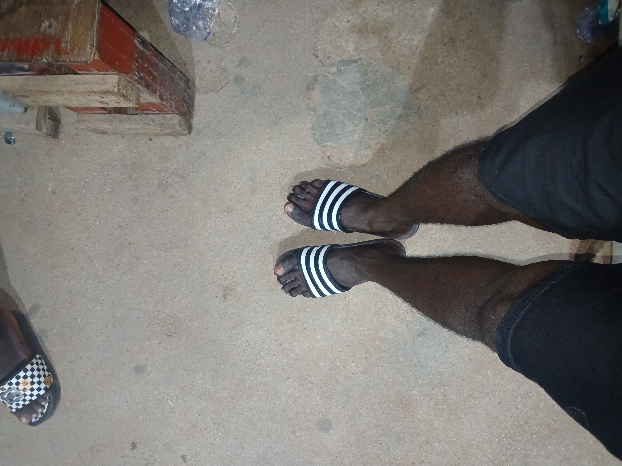 The image shows a close-up, high-angle view of a person's legs and feet. The person is wearing black shorts and black and white striped slide sandals. Their feet are bare and resting on a concrete or asphalt surface. In the background, to the left, another foot wearing a black and white checkered slide sandal is visible. To the right, there are some wooden planks and a plastic bottle. The scene appears to be outdoors, possibly in a public space within Jalingo, Nigeria, given the context provided, though specific landmarks are not identifiable. The lighting suggests it might be evening or under artificial light.