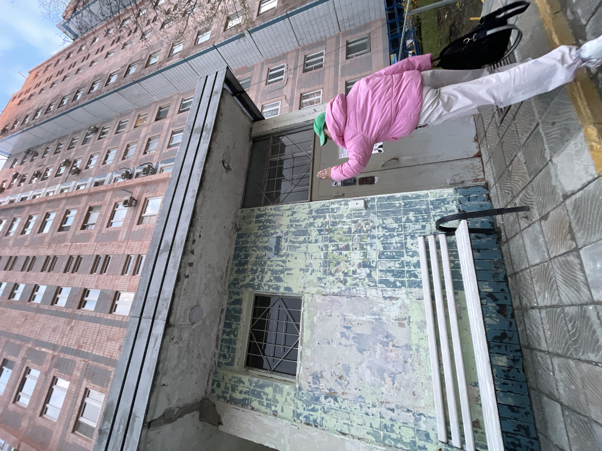 The image, captured from a low angle and severely rotated clockwise, shows a person entering a building, likely in Moscow, Russia. The individual, dressed in a bright pink puffer jacket, light-colored pants, and a green cap, is seen reaching out towards a grey entrance door. A black bag is slung near their hip.
The building is a tall, multi-story structure, possibly an apartment block or administrative building. Its facade features light brown brickwork interspersed with darker grey/blue tiled sections, containing many rectangular windows and several visible air conditioning units. The entrance area contrasts with the main facade, displaying heavily weathered and peeling blue and green textured tiles. A small window with decorative security grilles is located to the left of the entrance door.
The setting appears to be an older urban environment, characterized by the building's architecture and the worn state of the entrance. The sky is overcast, suggesting a cloudy day, and the person's warm clothing indicates cool weather. The activity depicts a routine action of accessing a building. No discernible text is visible on the building or door components.