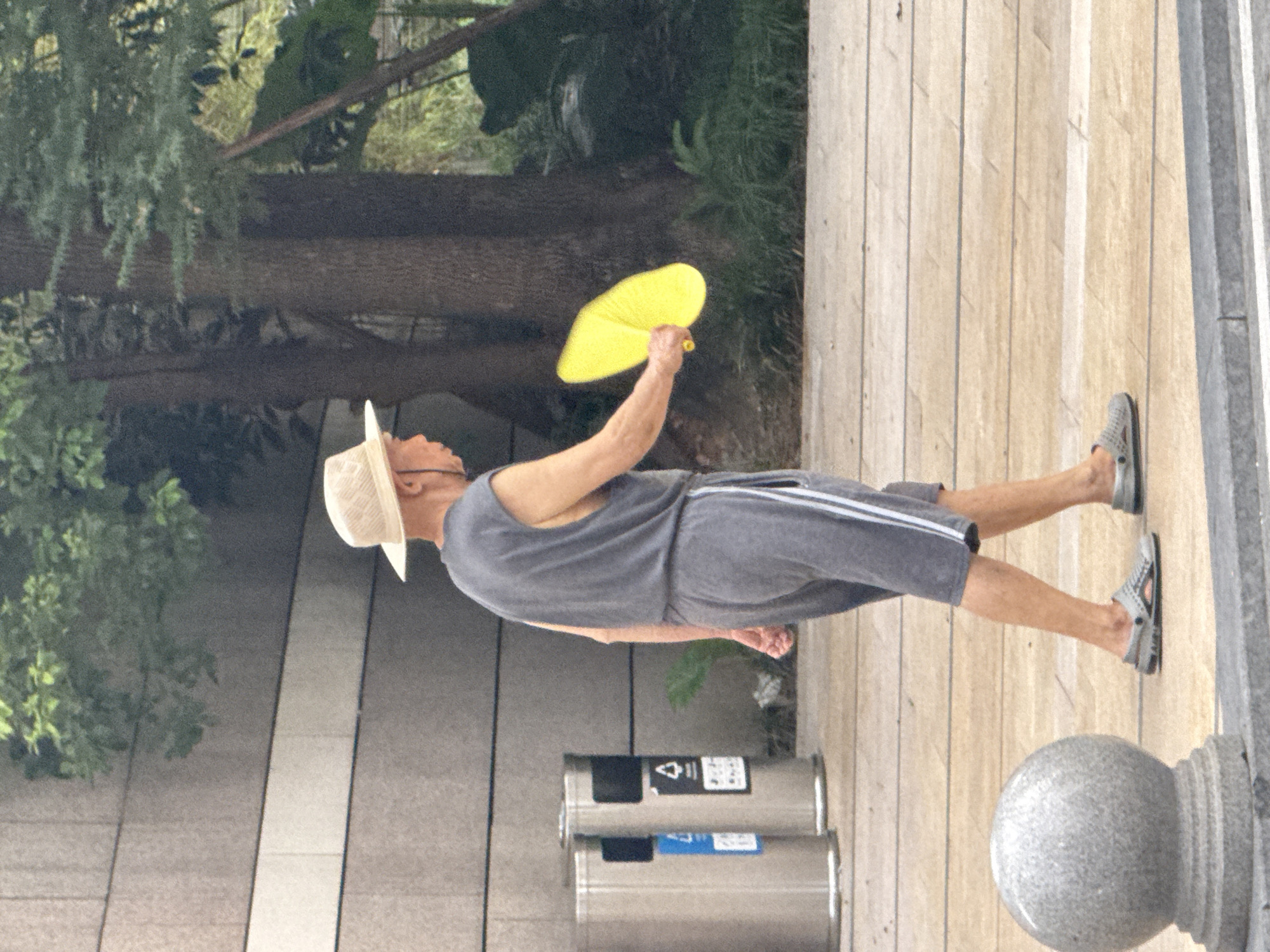 A full shot captures a person in profile, walking on a paved walkway near a building in Yuzhong District, China. The individual, wearing a straw hat, a sleeveless gray shirt, and gray shorts with white stripes, holds a bright yellow fan in their right hand. Two cylindrical trash cans are visible to the left of the person. The environment includes trees with dark trunks and green foliage, and a gray, spherical decorative element on a pedestal is positioned in the foreground to the left. The pavement is light-colored and tiled, with gray border stones visible at the bottom of the frame.