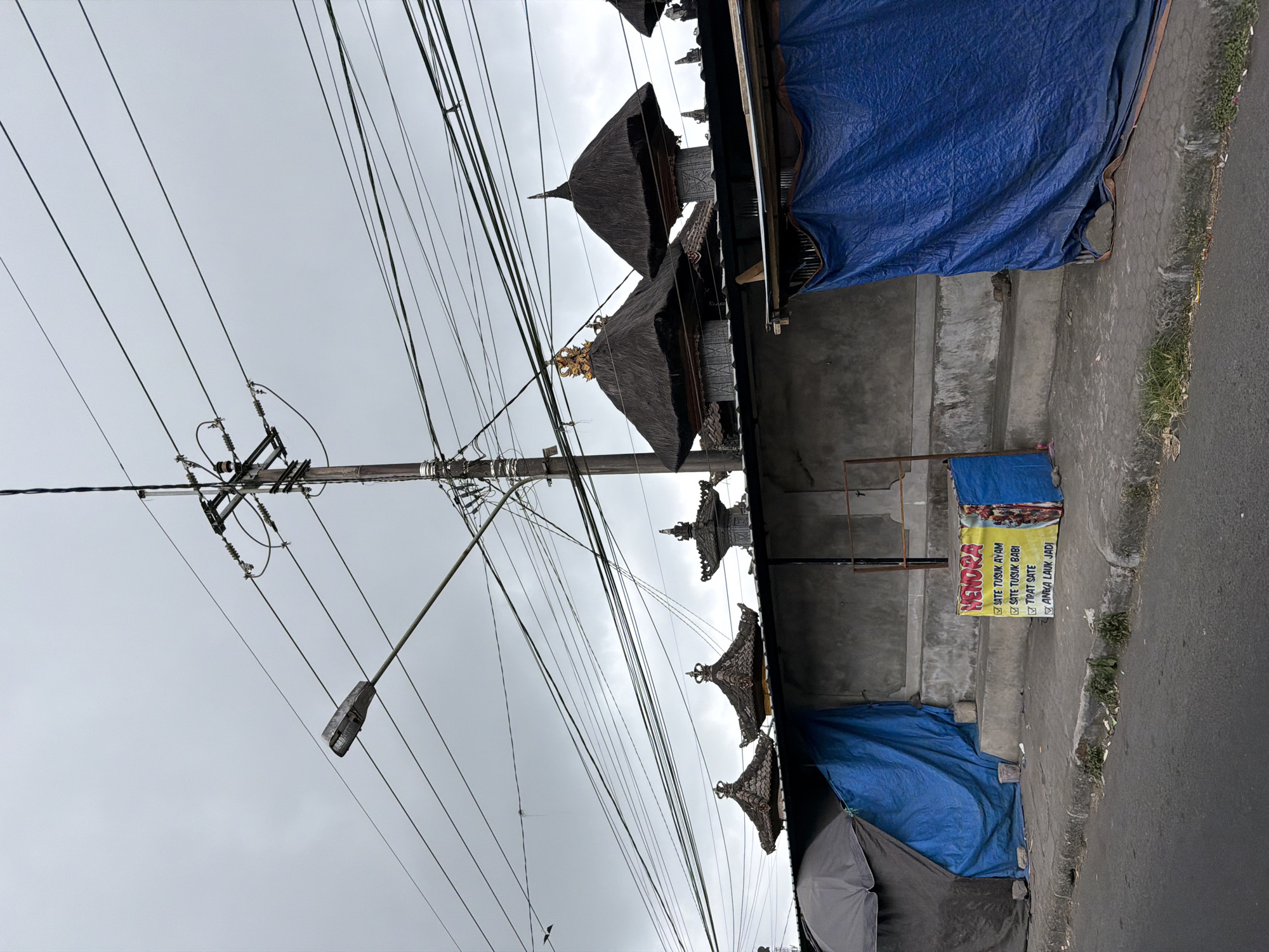 This media file captures an outdoor scene dominated by a dense network of overhead power lines crisscrossing the frame against a cloudy sky. Below the wires, a row of traditional thatched-roof structures with ornate, golden finials suggests a cultural or religious setting, possibly temples or pavilions, common in Kintamani, Indonesia. These structures are partially obscured by large, bright blue tarps that appear to be covering or protecting parts of the buildings or surrounding areas.
In the foreground, a concrete sidewalk or pathway runs alongside a road. A yellow sign is attached to a metal frame near the sidewalk. The sign displays text in Indonesian, reading "HENORA" at the top, followed by a list of food items with checkboxes: "SATE TUSUK AYAM," "SATE TUSUK BABI," "TIWAT SATE," and "AREM LAUK JADI." This indicates a food stall or eatery.
The overall impression is one of a somewhat utilitarian and weather-beaten street scene, where everyday infrastructure like power lines coexists with traditional architecture and local commerce. The overcast sky suggests it could be daytime, possibly morning or late afternoon, with no strong shadows visible. The presence of tarps might imply ongoing construction, protection from the elements, or temporary closures. The scene conveys a sense of everyday life in a location with distinct cultural elements.