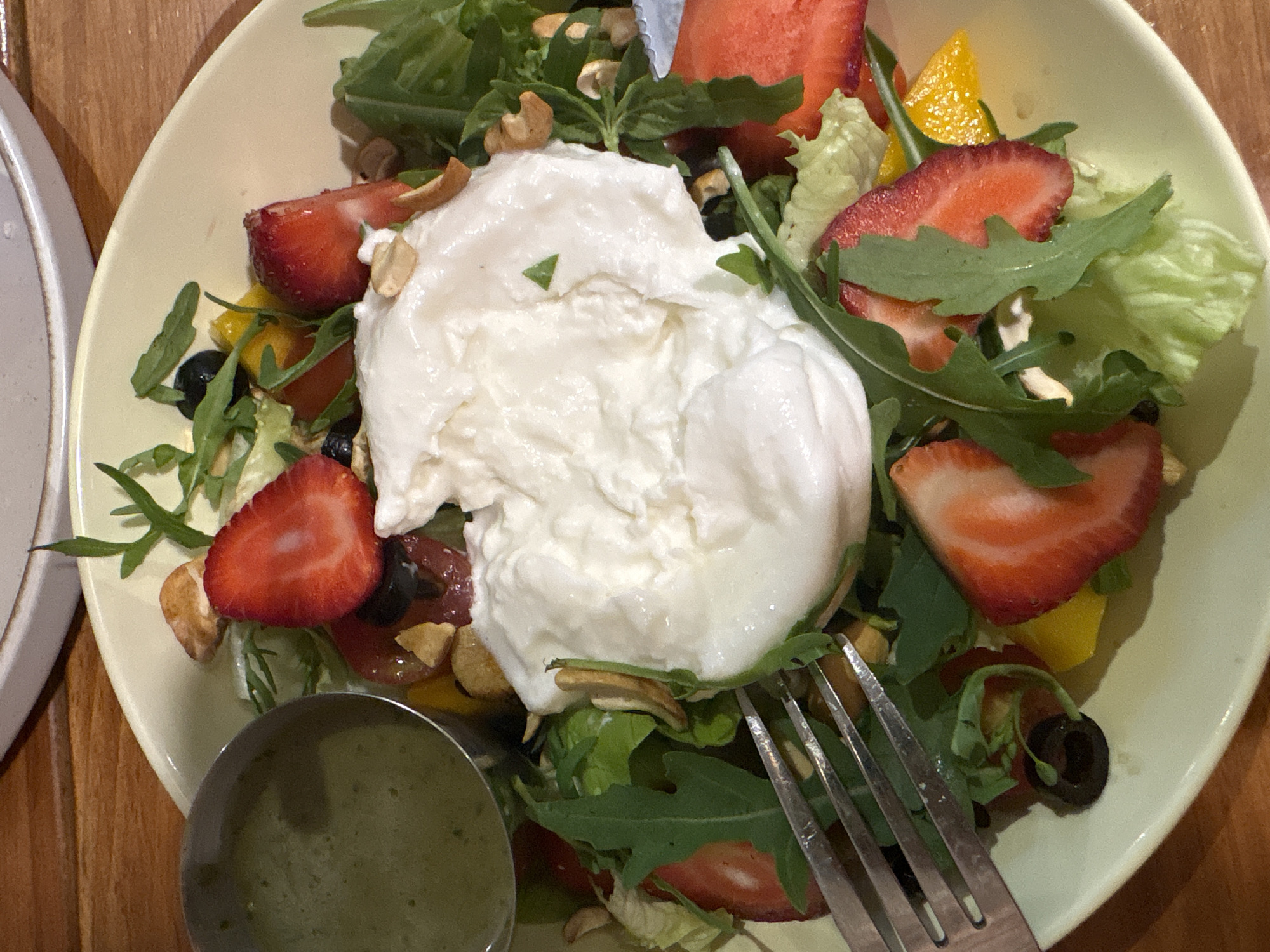 A close-up, overhead shot displays a salad served in a light green bowl on a wooden table. The salad features a large dollop of creamy white cheese, possibly burrata, at its center. Sliced strawberries, blueberries, black olives, and what appear to be nuts are scattered throughout the greens, which include arugula and lettuce. A small metal ramekin filled with a green dressing is positioned on the left side of the bowl. A fork rests on the left edge of the salad, partially submerged in the greens. The lighting suggests an indoor setting, possibly a restaurant. The specific location in Đà Nẵng, Vietnam is not determinable from the image.