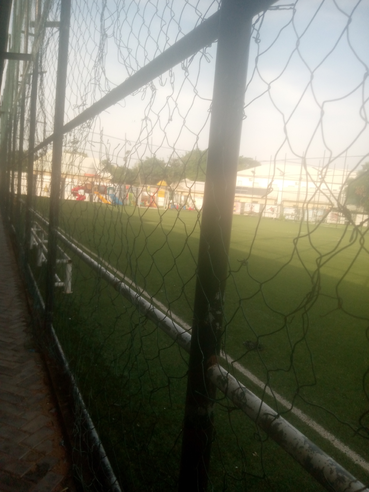 The image shows a view of a sports field, likely a soccer or football field, through a chain-link fence. The field appears to be made of artificial turf and has white lines marking boundaries. In the background, there is a playground with colorful slides and structures, suggesting the location is a school or community center. A building with a light-colored facade and possibly windows is visible behind the playground. The sky is bright, indicating it is likely daytime, perhaps late afternoon given the soft lighting. The fence in the foreground is somewhat worn and has some torn sections. A paved walkway or path is visible to the left of the fence. The overall scene suggests a recreational or educational facility in Jalingo, Nigeria. No people or specific activities are clearly visible, but the presence of the playground and sports field implies a place for children and sports enthusiasts. The lighting and hazy atmosphere suggest it might be a warm or humid day.