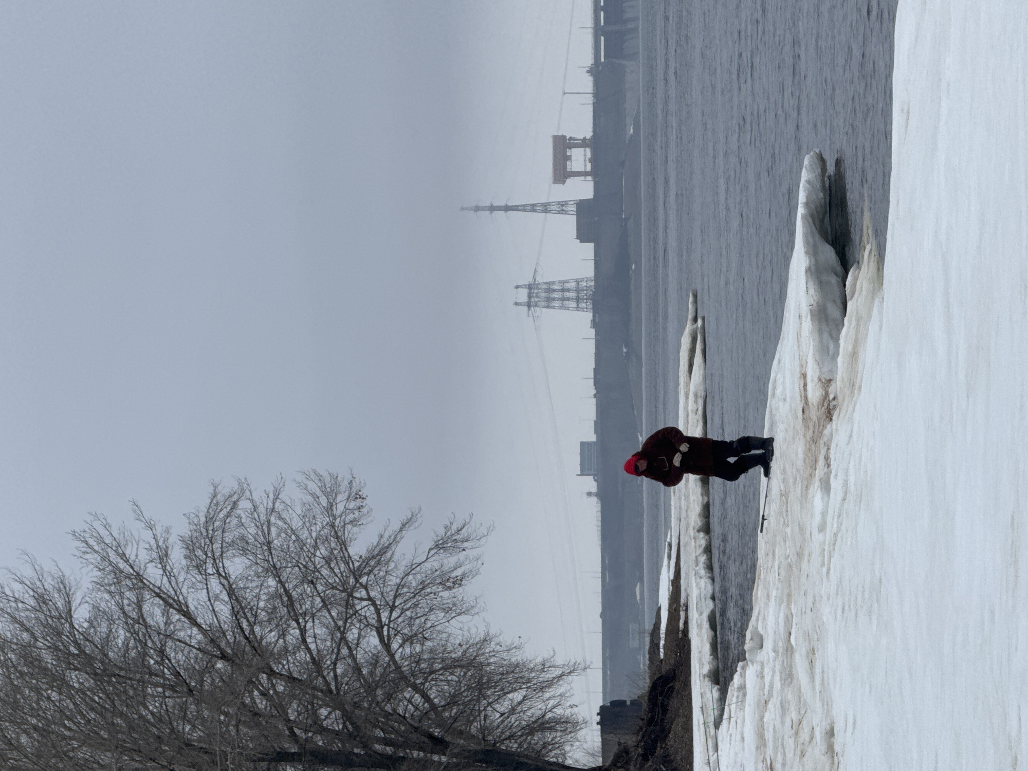 A person stands on a snow-covered bank near a body of water in Chaikovsky, Russia. The individual, wearing a red hat, dark jacket, and dark pants, faces away from the camera. The environment is overcast, and the background features a river or large body of water, as well as industrial structures including power lines, cranes and buildings on the opposite shore. A bare tree is visible in the foreground. The bank is composed of a dirt/snow mixture. The person has a hiking pole and has been captured in a walking motion.