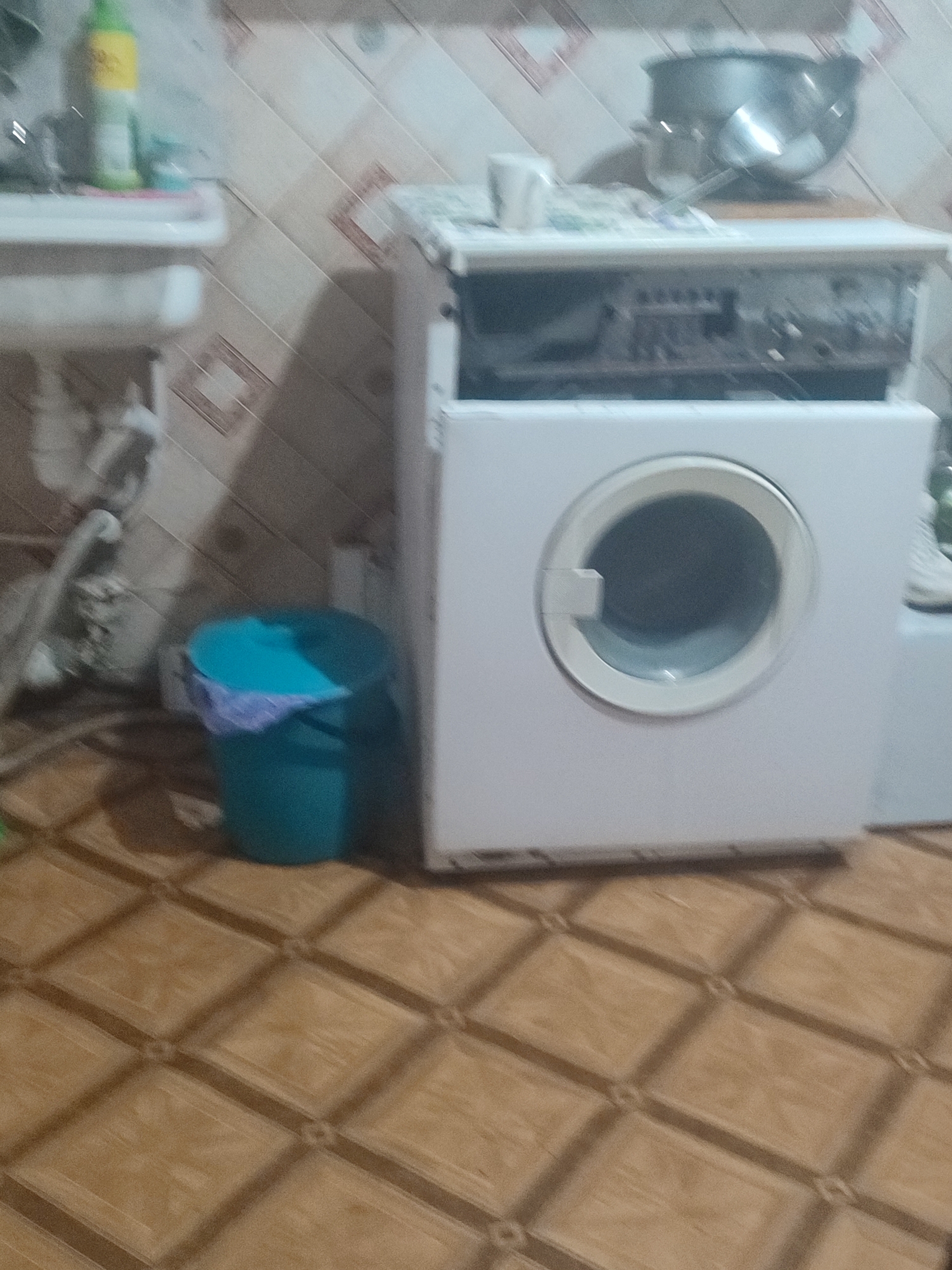 The image depicts a domestic scene, likely a utility area or laundry room, with a washing machine as the central object. The washing machine is white and front-loading, with a circular door. Above the washing machine, a part of what appears to be a stove or countertop is visible, with a large metal pot and a ladle on top. To the left of the washing machine, a white sink is partially visible, with a green bottle of cleaning product on its edge. A turquoise trash can with a crumpled cloth inside is situated to the left of the washing machine. The floor is tiled with a diamond pattern in shades of brown and beige. The walls are tiled with a pattern of beige squares with brown diamond accents. The overall lighting is dim, contributing to a slightly blurred and indistinct quality of the image. The scene is set in an interior space that is likely a residence. Based on the available information, the specific location within Chernivtsi, Ukraine cannot be determined from the visual cues alone.