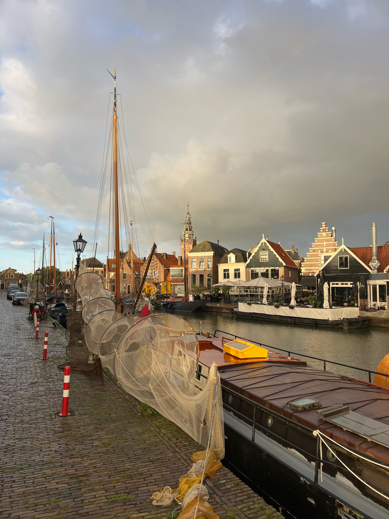 This is a photograph of the harbor in Monnickendam, Netherlands, taken from the cobblestone embankment. Several sailboats are docked along the canal, their masts reaching towards a cloudy sky. Fishing nets are draped over the sides of some of the boats. The embankment is lined with bollards and a street lamp. In the background, there are traditional Dutch houses with gabled roofs and a church tower. The scene suggests a calm, early morning or late afternoon, with the sunlight casting long shadows. There are no people visible in the image.