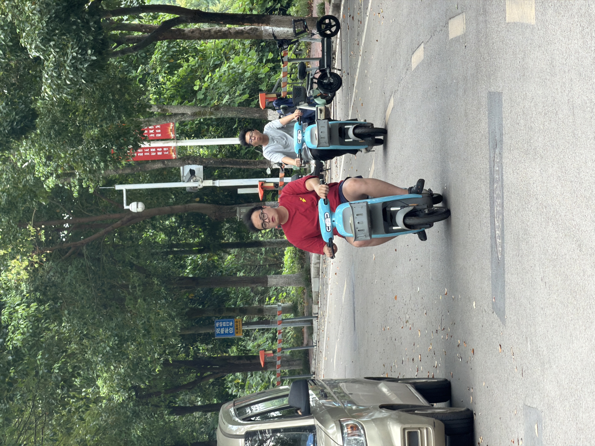 Two young men are riding electric scooters on a paved road lined with trees. The man in the foreground, wearing a red t-shirt and glasses, is riding a blue scooter. Behind him, a man in a grey t-shirt is riding another blue scooter. A gold van is parked on the left side of the road. Orange and white barriers are visible along the roadside, and a sign with Chinese characters is posted on a pole. The environment appears to be a park or residential area in Jiangjin, China.