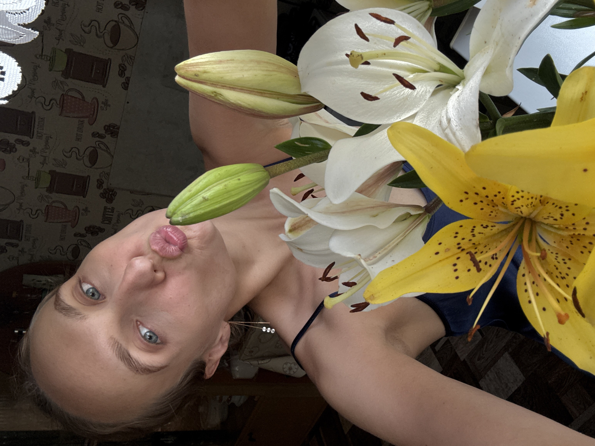 A young woman with blonde hair and blue eyes is taking a selfie in what appears to be a domestic setting, possibly in the city of Sharkan, Russia. She is holding a bouquet of lilies, predominantly yellow and white, in front of her face, with her lips pursed as if blowing a kiss. The background shows a patterned wallpaper and a piece of white lace. She is wearing a dark blue sleeveless top and a delicate earring with three small dangling stones. The lighting suggests it might be daytime indoors.