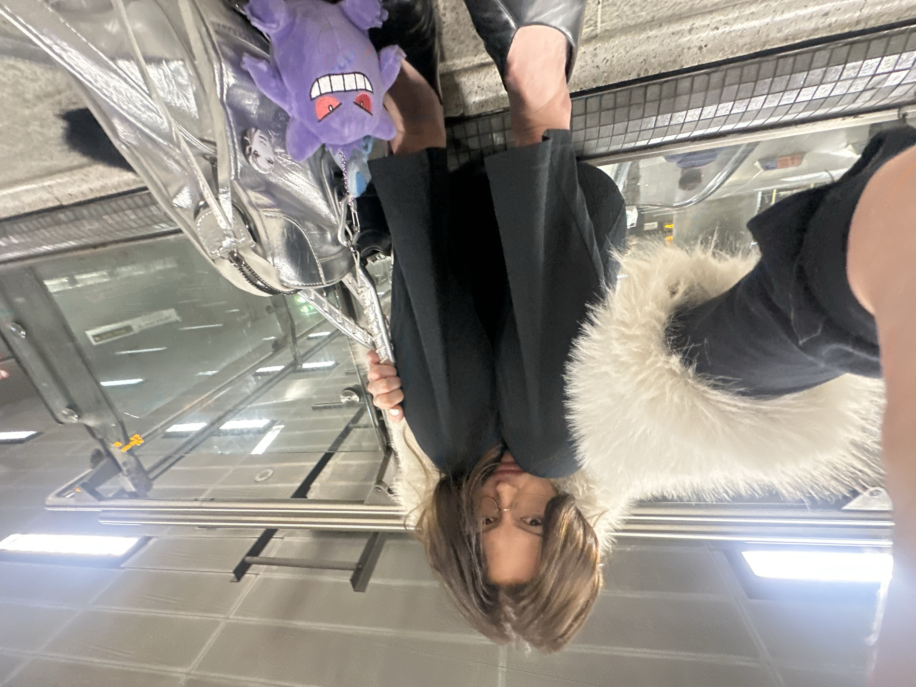 The image depicts an adult female situated on a stone ledge or step adjacent to an escalator within an underground station on the Piccadilly Line in London, United Kingdom, confirmed by a visible reflection of a "Piccadilly Line" sign. The subject, viewed from a low-angle, close-up perspective, has brown, shoulder-length hair and wears gold-rimmed glasses. She is dressed in a dark garment with a prominent, fluffy white or cream-colored faux fur collar. A metallic silver-colored handbag with a textured surface and chain-link straps rests beside her, held by her right hand, which exhibits white nail polish. Attached to the handbag are a purple Gengar plush toy with red eyes and a white-toothed grin, a smaller blue plush charm, and a monochromatic pin featuring a face. Her left leg is partially visible, and her visible foot wears a black flat shoe. The background includes a glass escalator barrier, another descending escalator with a blurred human figure, and a grey paneled ceiling illuminated by rectangular fluorescent light fixtures.