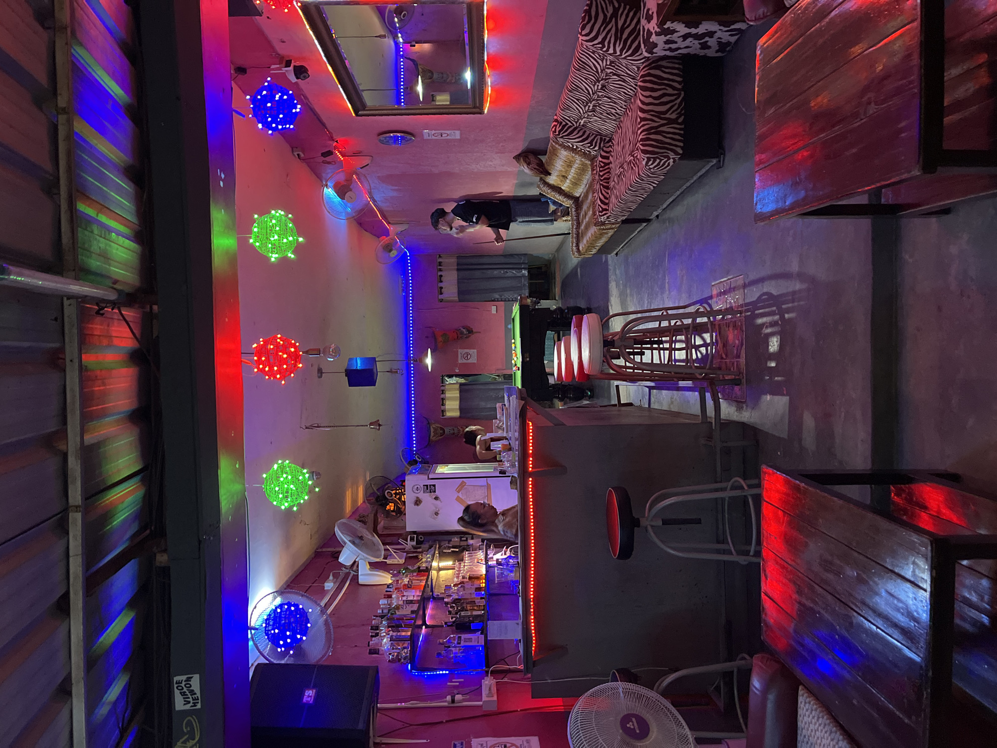 The image shows the interior of a bar or entertainment venue in Chiang Mai City Municipality, Thailand. The location is dimly lit with colorful neon lights and decorative spherical lights. A zebra-striped sofa is present, and a man is seen playing pool. A bar area is visible, with bottles displayed behind it and customers present. There are tables and stools, along with various fans.