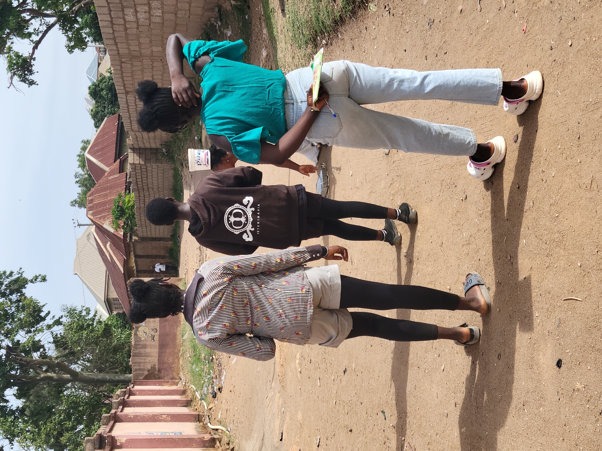The image depicts three young individuals standing on a dirt road in Jos, Nigeria. The environment includes modest buildings with rusted roofs, visible garbage, and rudimentary infrastructure like a cement stairway. The individual nearest to the viewer wears a striped shirt, shorts over black leggings, and slides. The middle person has on a brown sweatshirt and black leggings, and the individual farthest away wears a turquoise top and light-colored jeans with white clogs. The image suggests a residential area with limited development, indicating a likely low-income neighborhood.