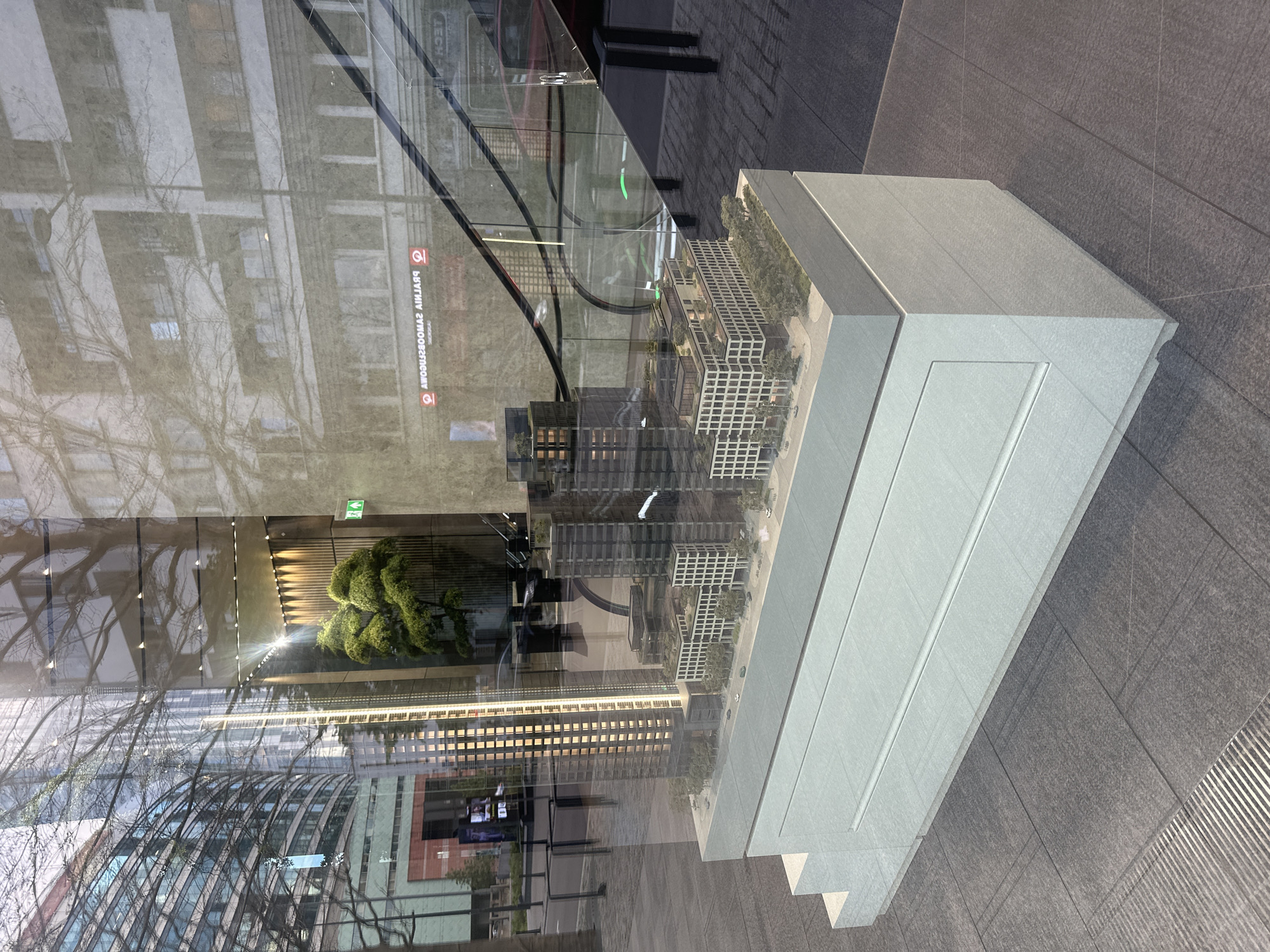 This is a photograph of an architectural model of a modern building complex, displayed behind a glass window in a city. The model features several high-rise buildings and lower-rise structures with detailed facades, greenery, and miniature vehicles. The model is situated on a multi-tiered white base, which rests on a tiled floor.
The reflection in the window provides context about the surrounding urban environment. On the left side of the reflection, a large, modern glass-fronted building with multiple stories is visible. In the center of the reflection, a bare tree with intricate branches is prominent. To the right, an escalator is visible, indicating an indoor public space or a building entrance. Above the escalator, text is reflected, which reads "PRALNIA BOOM AWAJJAQ" and "TECH-H" which are reversed, suggesting it is written on a sign outside the window. This reversed text, specifically "PRALNIA BOOM AWAJJAQ", can be deciphered as "QAJAWA MOOB MAJNALRP" which is not a coherent phrase. However, considering the reflected text "TECH-H" which is likely "H-TECH", it is probable that the other text is also reversed and potentially part of a company name or slogan.
The presence of the text "TECH-H" and the architecture of the buildings in the reflection, combined with the style of the architectural model, strongly suggests the location is Warsaw, Poland. Specifically, the architectural style and the type of development modeled are consistent with modern urban regeneration projects often seen in Warsaw. The reflection of an escalator and the commercial-looking signage points to a commercial or residential development in a busy urban area. The overall scene depicts a display of a new property development. There are no people clearly visible in the immediate scene, only reflections of the urban environment.