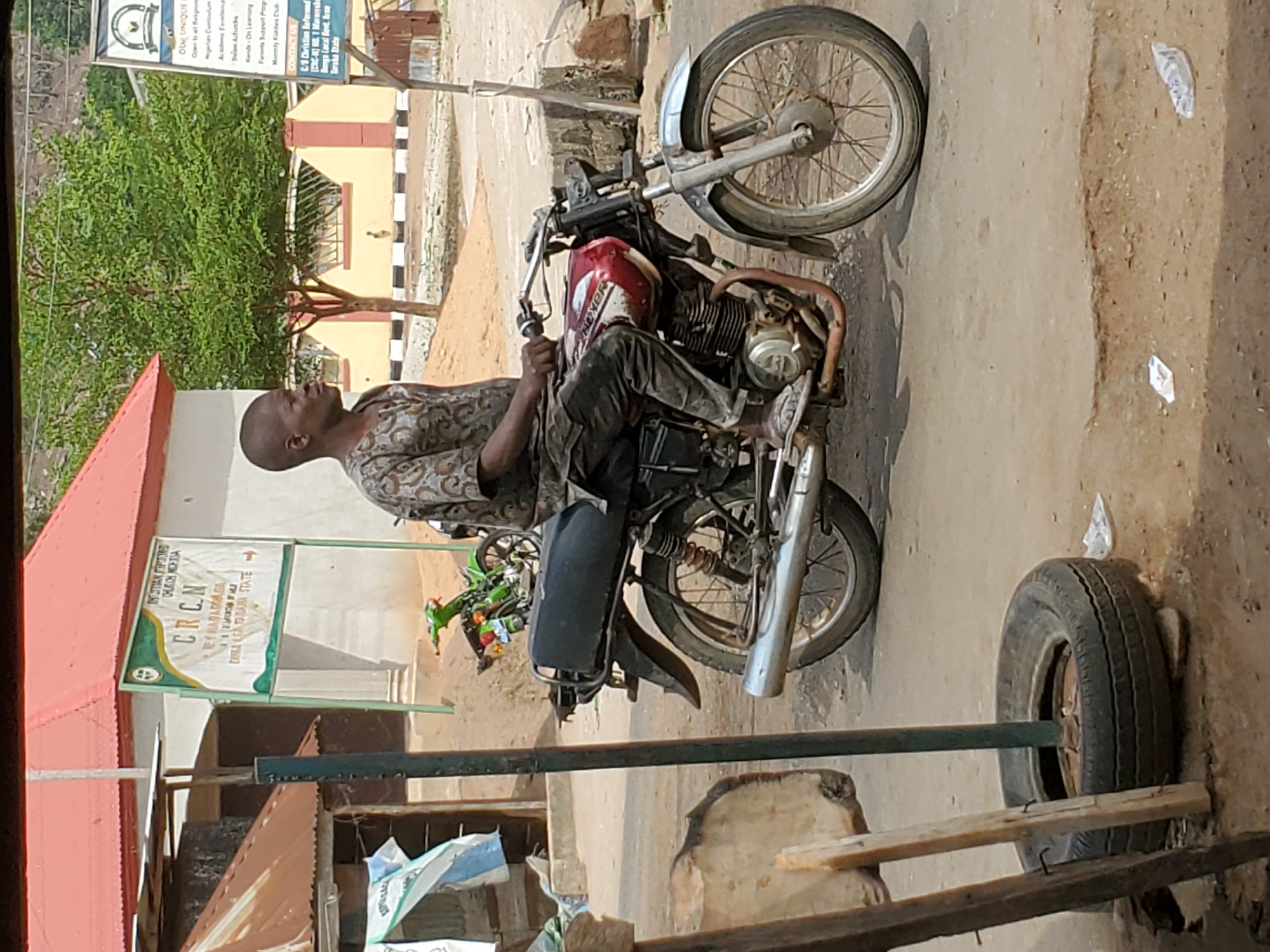 A medium-shot, outdoor photograph shows a man seated on a Boxer-brand motorcycle in Mararaba, Nigeria. The man, bald and in profile, wears a patterned, earth-toned shirt and dark trousers. He sits on the motorcycle, which has a red tank and is parked on a concrete surface. A spare tire leans against a wooden structure nearby. A sign with green and white lettering, partially obscured, identifies the location as "CRCN Central Reign Church Mararaba, Danka LGA Taraba State." The background includes a beige building and green trees.