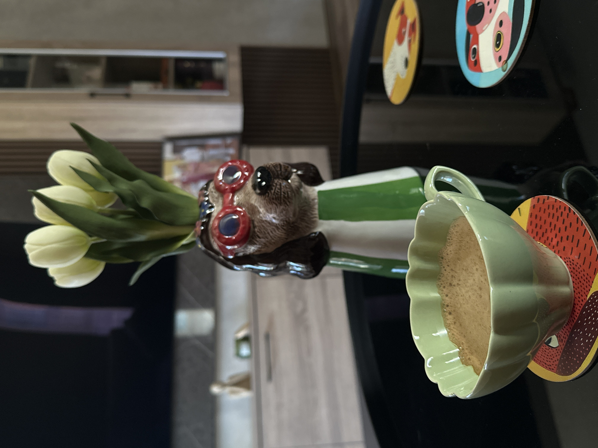 A close-up shot captures a light green scalloped cup filled with frothy coffee, placed on a red coaster with a patterned design, both resting on a glossy black table. To the left and slightly behind the cup is a ceramic vase shaped like a dog's head wearing red-rimmed glasses, holding a bouquet of white tulips with green stems and leaves. The background features a blurred living room setting with wooden cabinets and shelves, a television, and a framed picture. The image appears to be taken indoors, likely in a residential setting in Sofia, Bulgaria, given the context provided.