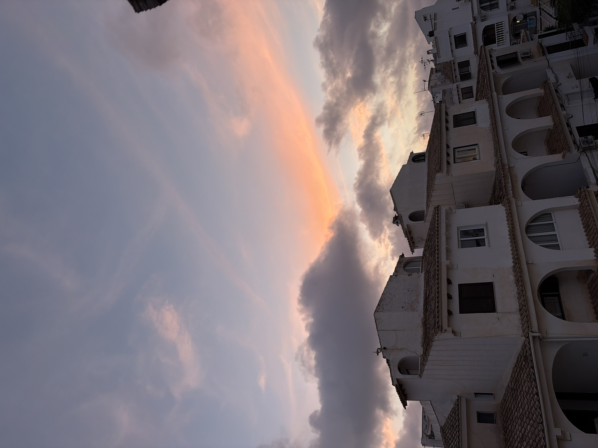 This media file depicts a building in Torrevieja, Spain, against a backdrop of a sunset sky. The building is white with multiple stories, featuring balconies with arched openings and tiled roofs. Various windows are visible, some with shutters. The sky displays a gradient of colors, with soft pinks and oranges from the setting sun blending into darker grays and blues of the clouds. The scene suggests a tranquil evening. No people are visible in the frame. No text is discernible. The image is oriented vertically.
