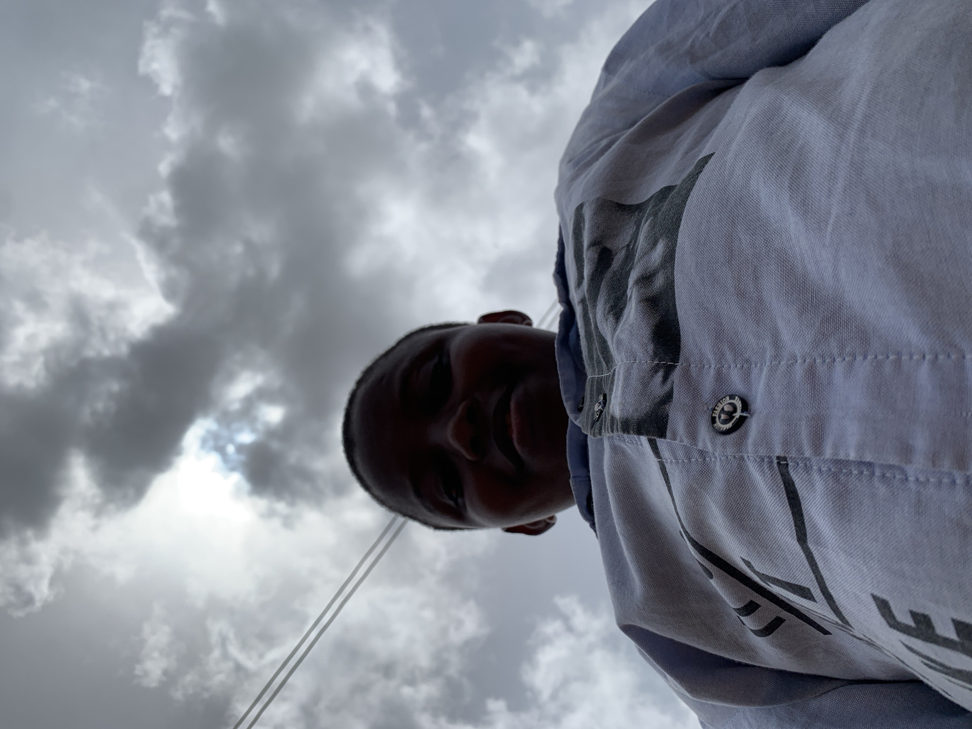 A low-angle shot shows a person with dark skin wearing a light-colored jacket against a backdrop of cloudy sky. The person is slightly out of focus, but they appear to be smiling. The sky is overcast with dark gray clouds and a small bright area indicating the sun. Based on the appearance of the clouds and sky, this scene can be assumed to be in Ikorodu, Nigeria, due to the prevalence of tropical weather patterns that are associated with this area. However, there are no specific details in the picture to explicitly confirm this. The focus is on the person and the sky.