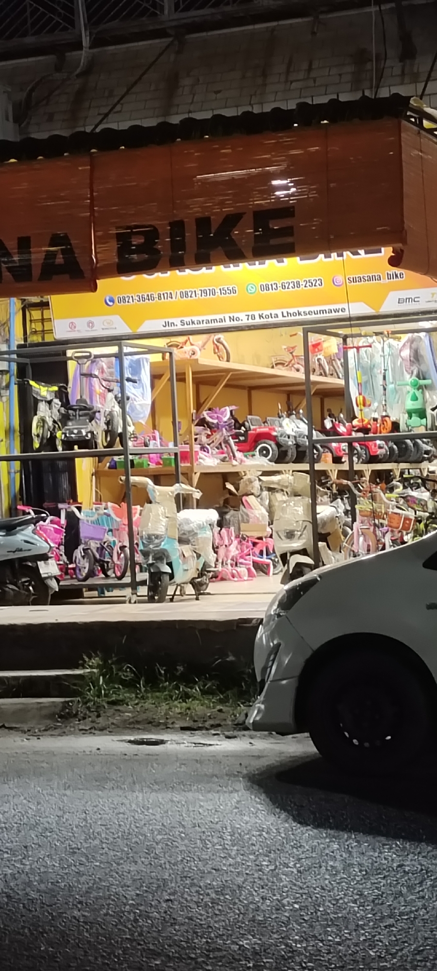 The image captures a brightly lit storefront at night, likely a bicycle and children's toy shop named "Suasana Bike," located in Lhokseumawe, Indonesia. The store's interior is visible, showcasing numerous children's bicycles, tricycles, ride-on cars, jeeps, and scooters, many still wrapped in protective plastic or packaging. These items are displayed on shelves and on the floor, indicating a wide variety of stock.
Above the store entrance, a yellow banner-style sign with black text is prominent. Parts of the store name "SUASANA BIKE" are visible, with "NA BIKE" being clearly legible. The sign also displays multiple phone numbers: "0821-3646-8174 / 0821-7970-1556" and "0813-6238-2523." An Instagram handle, "suasana_bike," is also present. Crucially, the address "Jln. Sukaramai No. 70 Kota Lhokseumawe" is clearly stated, confirming the location. A small "BMC" logo is also partially visible on the right side of the sign. Above the sign, a set of brown, bamboo-style blinds is rolled up. The building's upper facade features a light-colored brick wall.
In the foreground, a white car is partially visible on the right, with its front bumper and tire in the frame. The scene includes a paved road surface in front of the store, separated by a curb and a small strip of grass. A light-blue scooter, still largely covered in plastic, is parked on the store's elevated platform, and a small cat is visible near its wheels. The overall impression is that of a bustling local business, even at night.