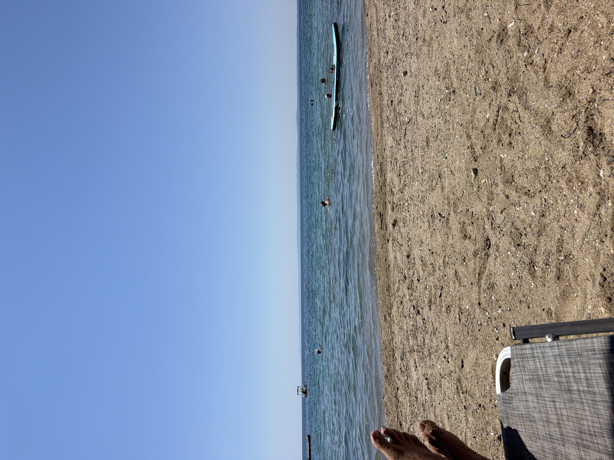 The image depicts a tranquil beach scene in Kalyves Polygyrou, Greece. The foreground features a sandy beach with a beach chair in the lower left corner, partially obscuring a person's feet. The sea stretches out to the horizon under a clear blue sky. In the water, several individuals are visible swimming or relaxing. One person appears to be on a paddleboard, which is a light blue color. Further out in the sea, a small boat is anchored. The overall atmosphere is one of leisure and summer enjoyment.