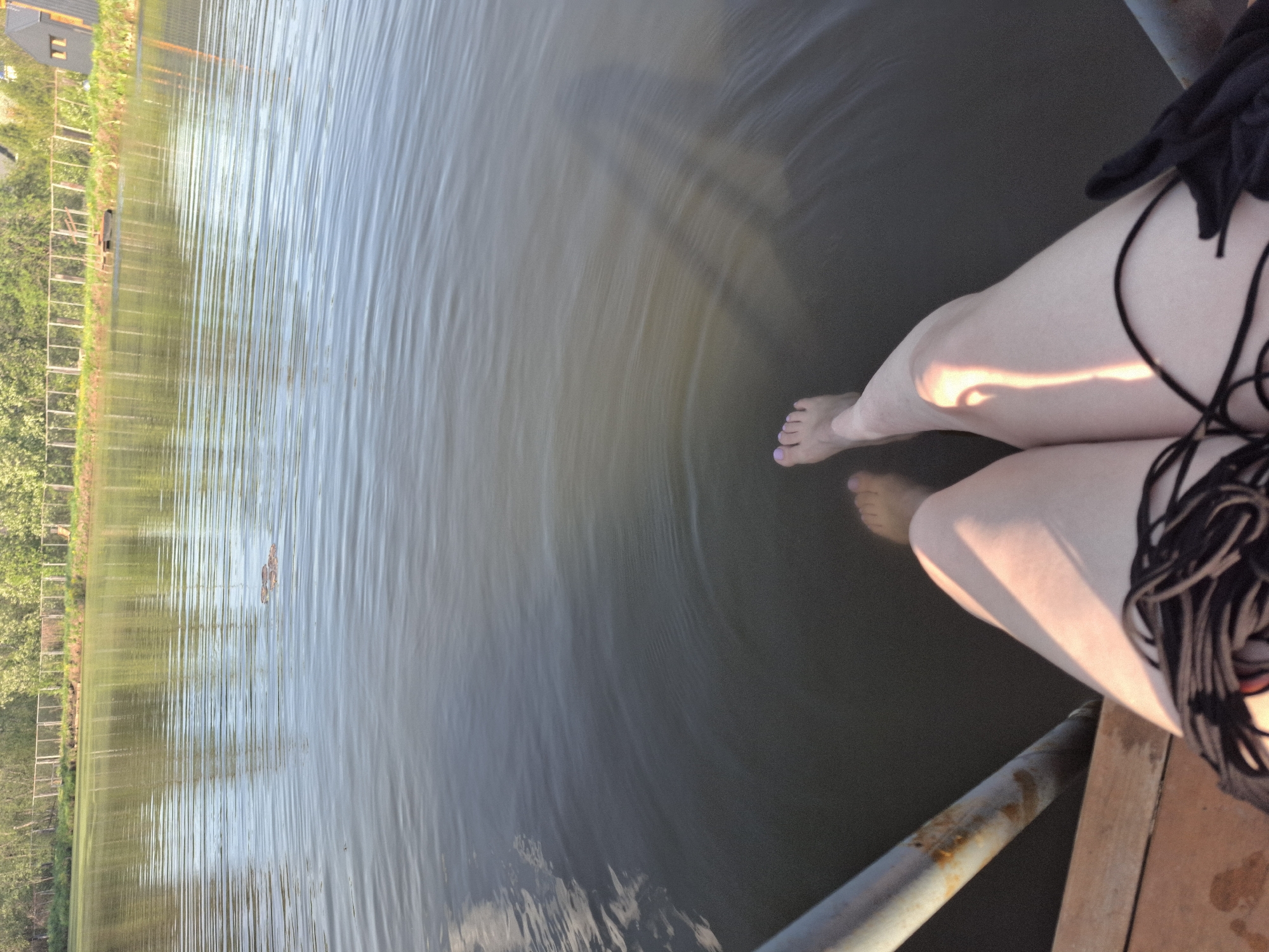 A view from a wooden dock or pier in Malyi Bukor, Russia, shows a person seated with their legs dangling into the water. The water is dark, reflecting light and ripples from the person's feet. The person has painted their toenails a light pink color. The lower half of the person’s body is positioned on the right side of the image; part of a black bathing suit is visible. A rusty metal railing is partially visible at the bottom edge of the image. Across the water, there is a green treeline and a few buildings.
