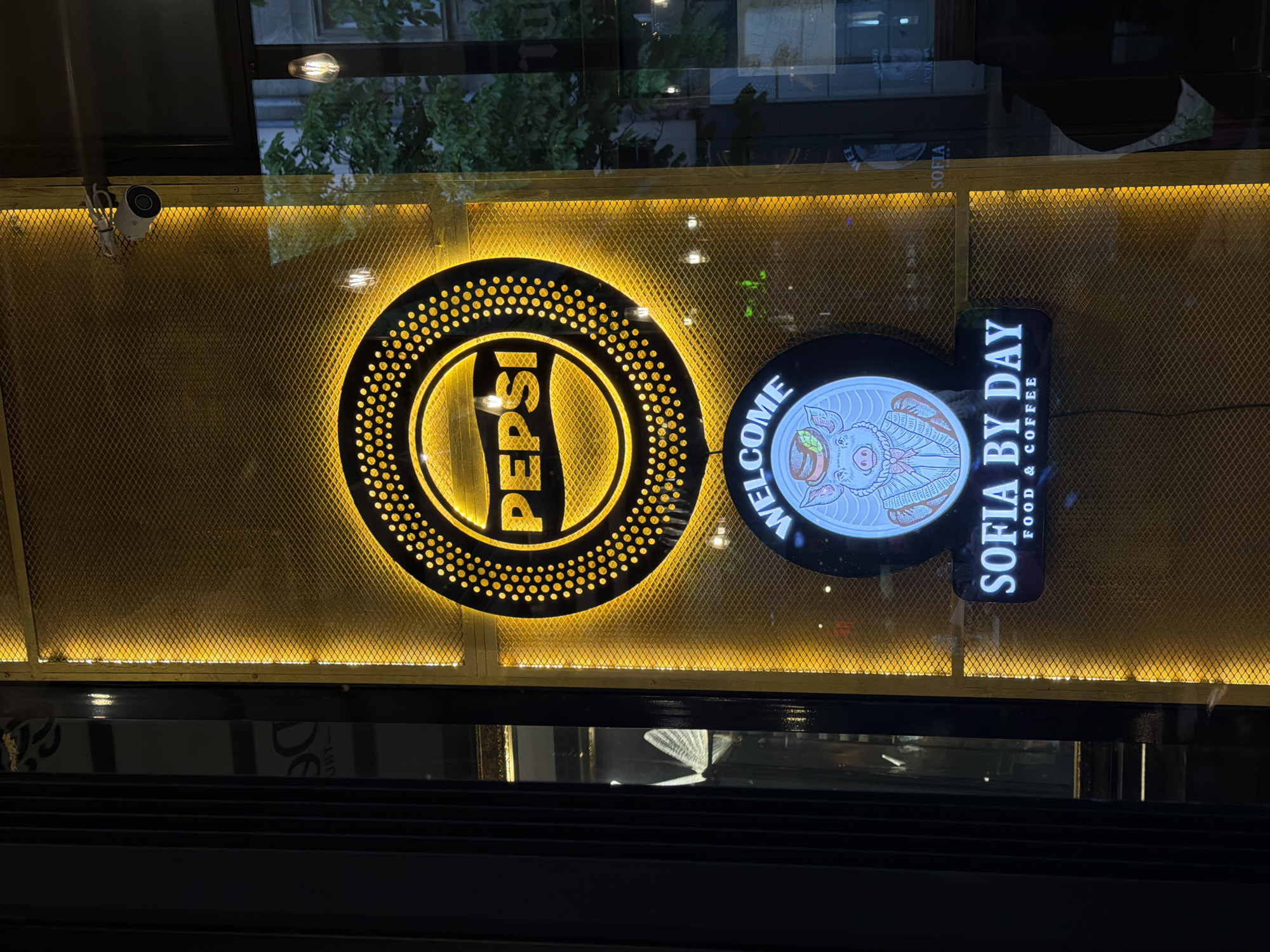 The image captures the illuminated storefront of a commercial establishment at night. Two distinct, glowing signs are prominently displayed through a reflective window.
On the left, a large circular sign features the iconic "PEPSI" logo in its characteristic red, white, and blue, though rendered in a golden-yellow light that bathes the word. The circular perimeter of this sign is adorned with numerous small, warm yellow lights, creating a sparkling effect.
To the right, another circular sign bears the text "WELCOME" curved across its top edge. The central part of this sign illustrates a stylized pig, possibly wearing a chef's hat or a leaf, against a light background. Attached vertically to the right side of this circular sign is a rectangular panel that reads "SOFIA BY DAY" in bold letters, with "FOOD & COFFEE" written in smaller text below it.
The background behind both signs is a textured, possibly metallic mesh, uniformly lit from behind with a warm, golden-yellow glow, contributing to the inviting atmosphere. Reflections of trees and exterior lighting are visible on the window glass, indicating an outdoor setting after dark. A small, white security camera is mounted in the upper left corner of the scene, above the mesh and near the top edge of the window. The lower portion of the image is dark, showing the base of the window or a dark surface below it.
The visible text "SOFIA BY DAY FOOD & COFFEE" strongly suggests the establishment is a cafe or restaurant named "Sofia By Day," located in Sofia, Bulgaria, as indicated by the context. The presence of the Pepsi logo implies it serves beverages from that brand. The overall impression is a vibrant and welcoming commercial space, illuminated to attract customers during nighttime hours.