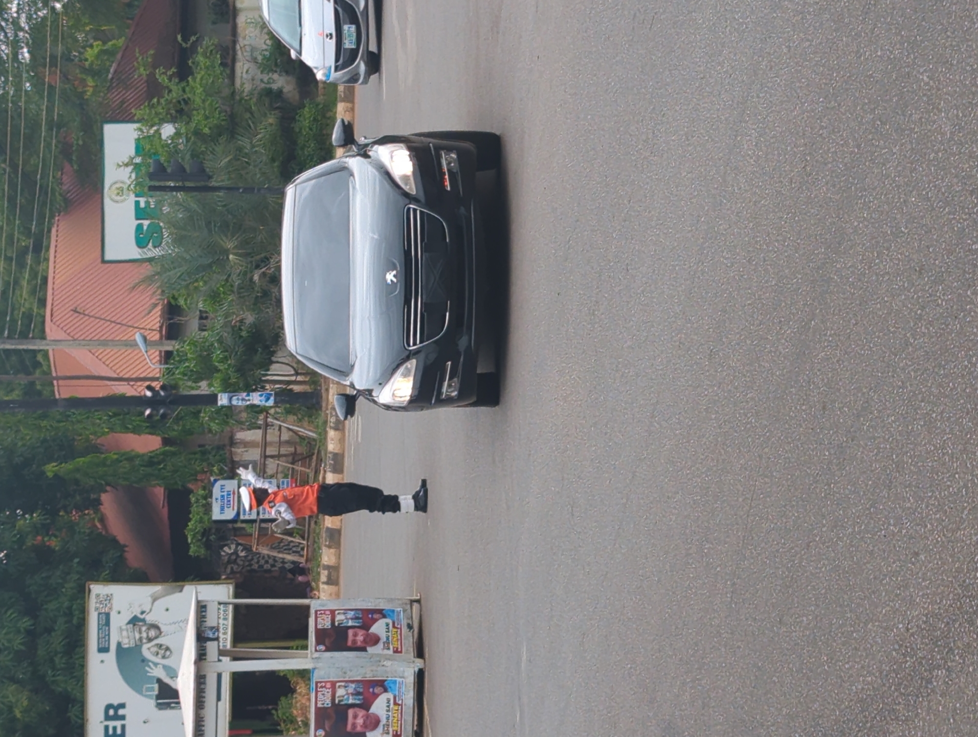 The image shows a black Peugeot car traveling down a street. A uniformed traffic officer is directing traffic. Political campaign posters are visible. A sign for "Thelish Eye Centre" can be seen in the background. There is also a sign for "SER." This appears to be in Shaba, Nigeria. Other cars are on the street. The environment includes trees and buildings.