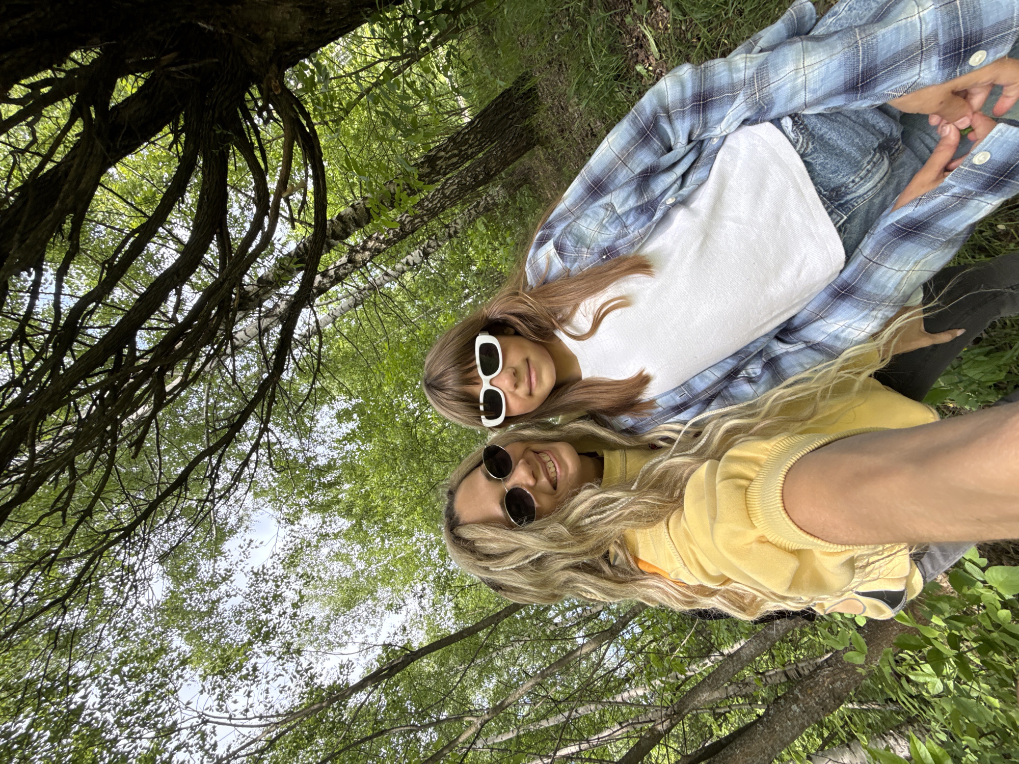 Two young women are outdoors in a wooded area. One woman wears a yellow sweatshirt and sunglasses; the other wears a white t-shirt, a blue plaid shirt, jeans, sunglasses, and black leggings. Branches of trees with green foliage fill the background and parts of the foreground.