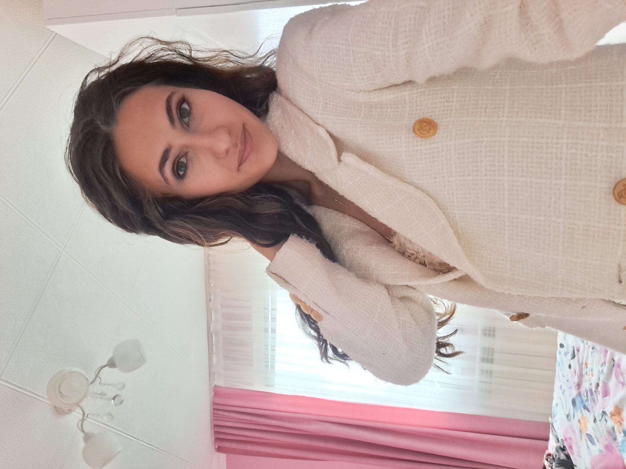 A woman with long, dark, wavy hair is taking a selfie indoors. She is wearing a cream-colored tweed blazer with gold buttons over a lacy pink top. Her right arm is bent, and her hand is touching her hair. The background shows a ceiling with a white light fixture, pink curtains, and sheer white curtains covering a window. A bed with a floral comforter is visible on the left side of the frame. The lighting suggests it is daytime, possibly with natural light coming through the window. The scene appears to be a bedroom or a living area. The image was taken in Negovan, Bulgaria.